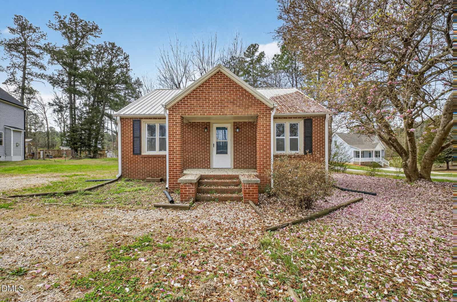 A small brick house with a metal roof, two front windows, a central door with steps, and a porch sits quietly at 5182 N Carolina 56 in Franklinton. The yard is dotted with scattered pink petals, and there are trees and neighboring houses in the background.