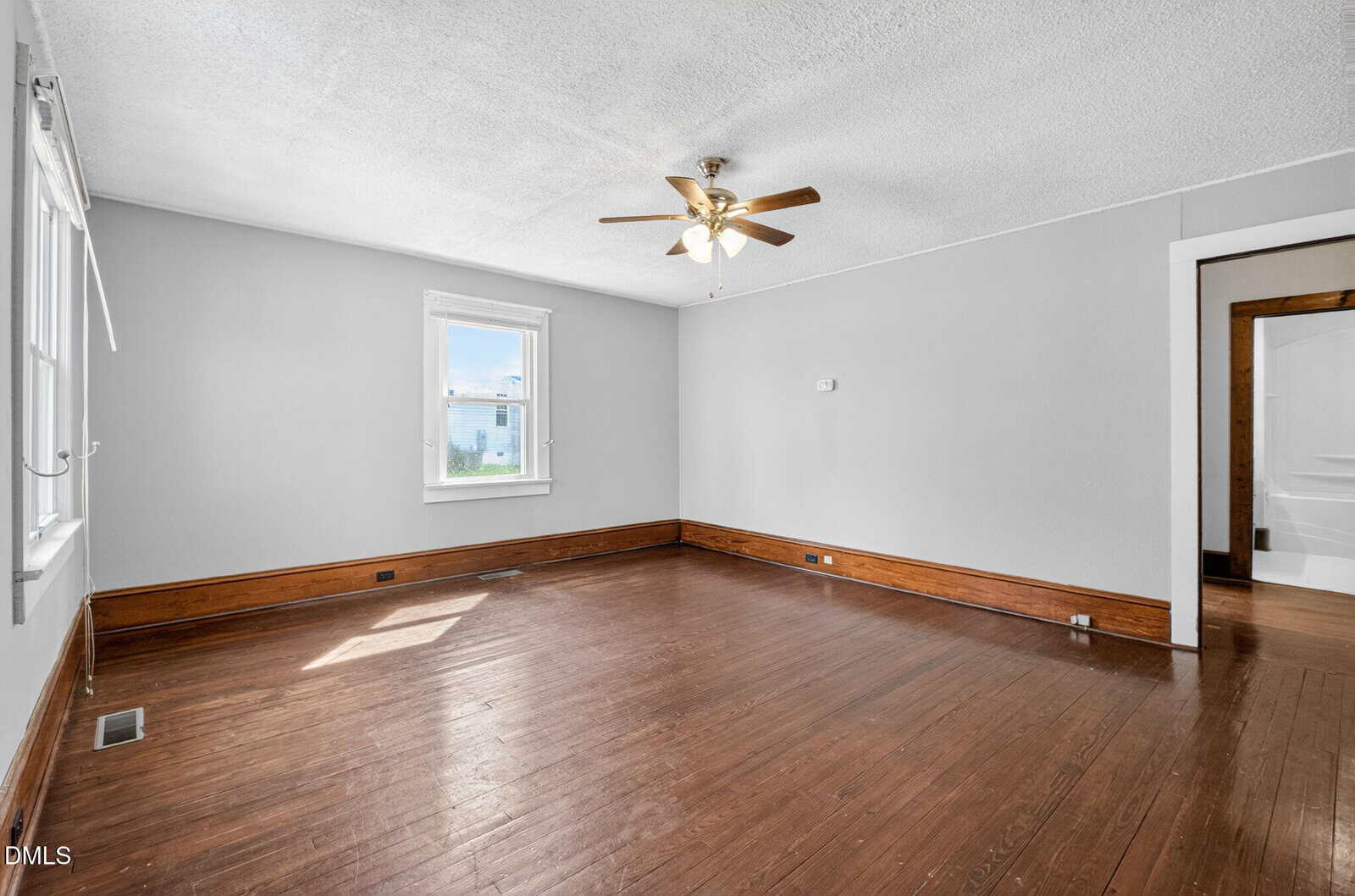 Empty room with light gray walls, a white ceiling, dark wood trim, and hardwood flooring. A ceiling fan with lights hangs in the center. Sunlight enters through two windows. An open doorway leads to a white-tiled bathroom.