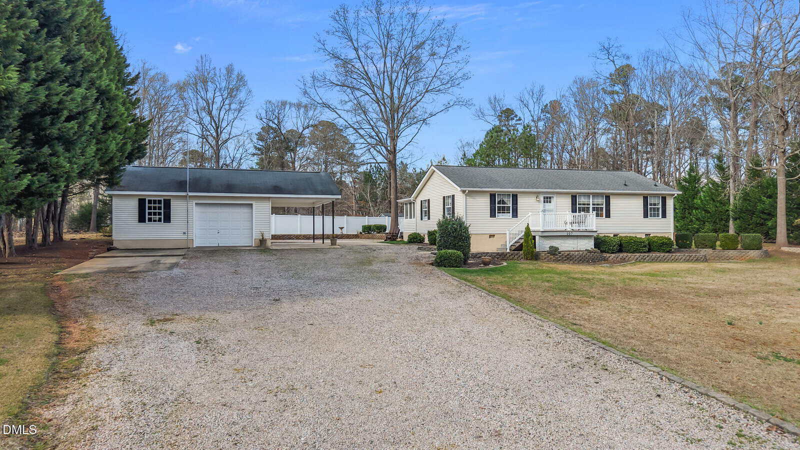 Single-story beige house with white trim and a front porch, located next to a detached two-car garage. There is a gravel driveway, sparse grass, and trees in the background under a blue sky.