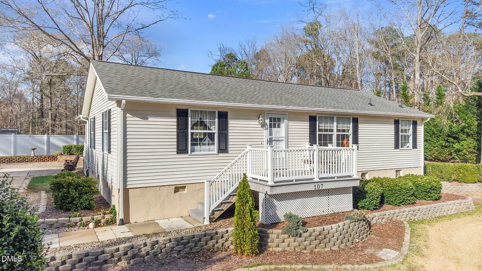 Single-story beige house with black shutters, white trim, and a small front porch. The yard has shrubs, a stone retaining wall, and landscaping. Trees and a blue sky are visible in the background.