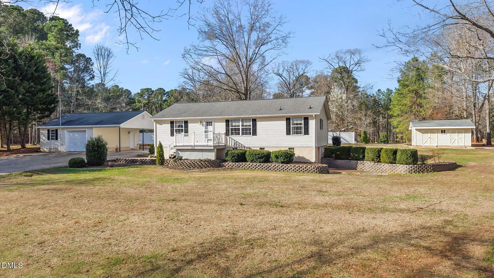 A single-story white house at 107 Blackfoot Drive, Louisburg sits on a raised foundation with a detached garage and small shed nestled in a spacious yard, surrounded by trimmed bushes and trees in the background under a clear blue sky.
