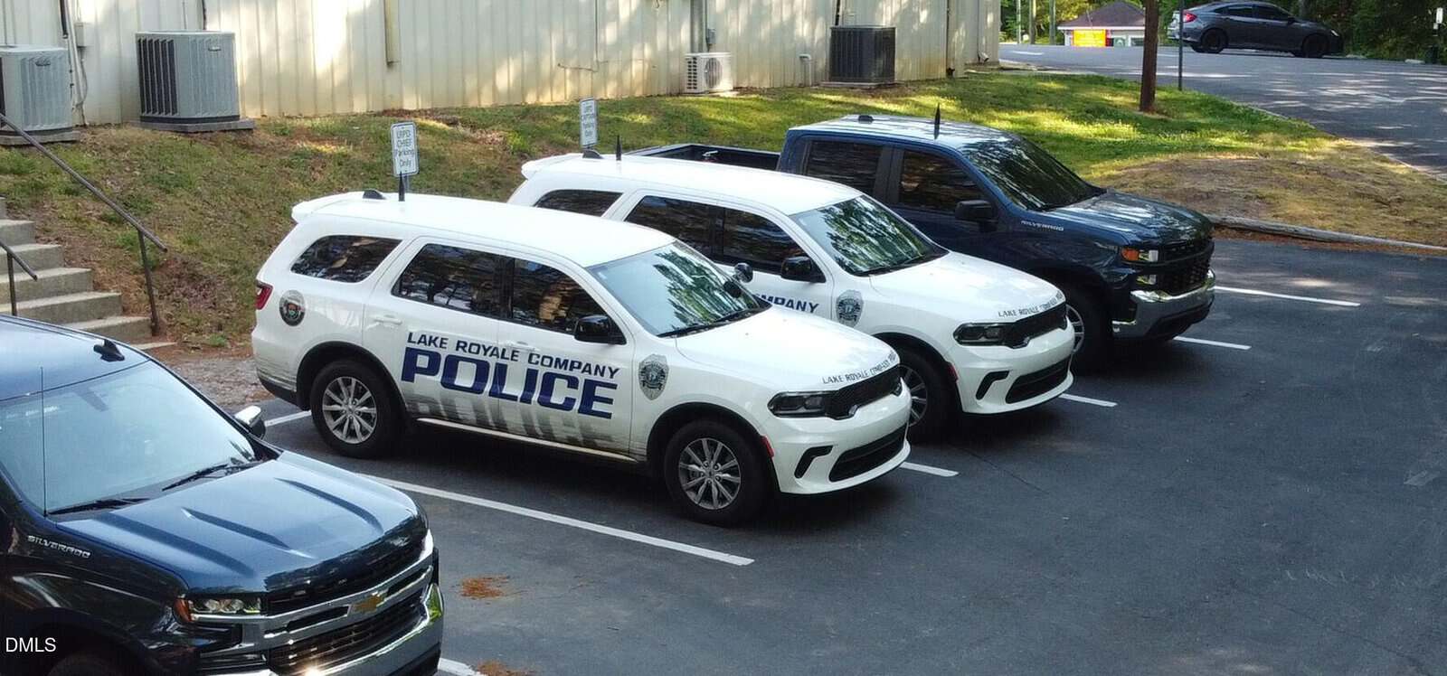 Three marked “Lake Royale Company Police” SUVs and one unmarked dark pickup truck are parked in a row in a paved parking lot near a beige building and some trees.