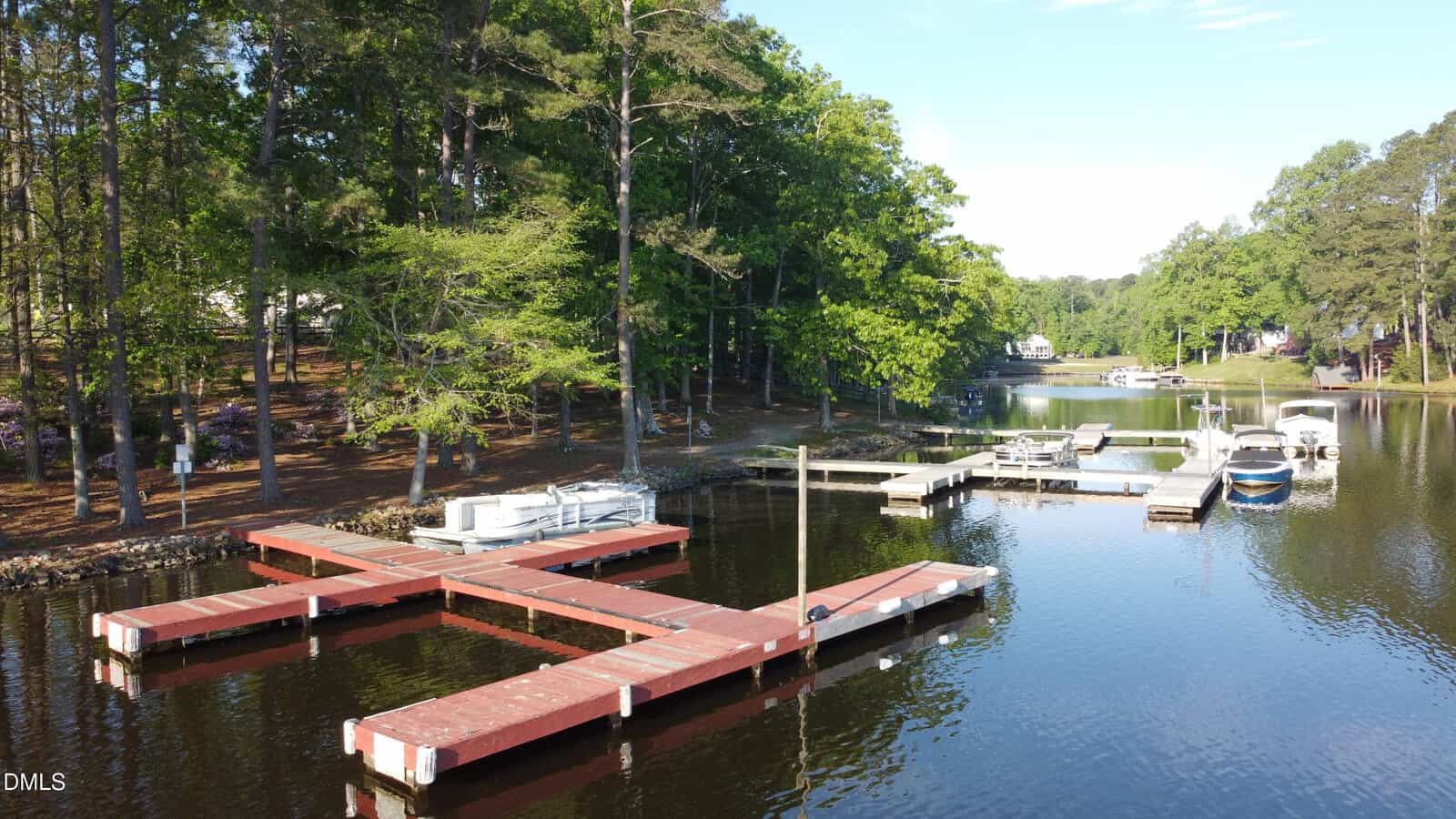 Red floating docks extend into a tree-lined lake. Several boats are tied to the docks. The water is calm, reflecting the surrounding trees. More docks and boats are visible in the background along the lakeshore.