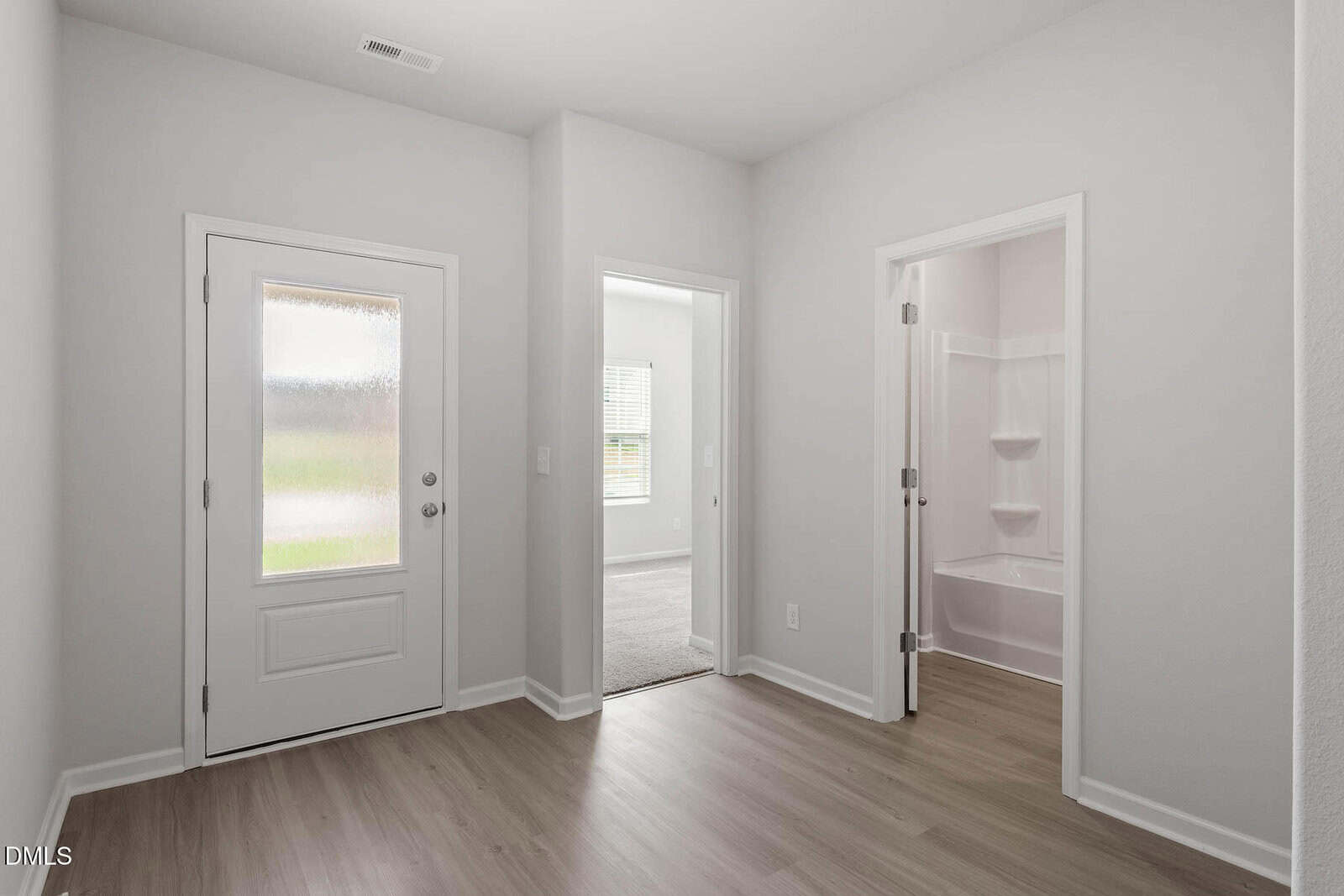 A bright hallway with light wood floors, a white door with frosted glass, and two open doorways leading to a carpeted room and a bathroom with a white bathtub and shelving.