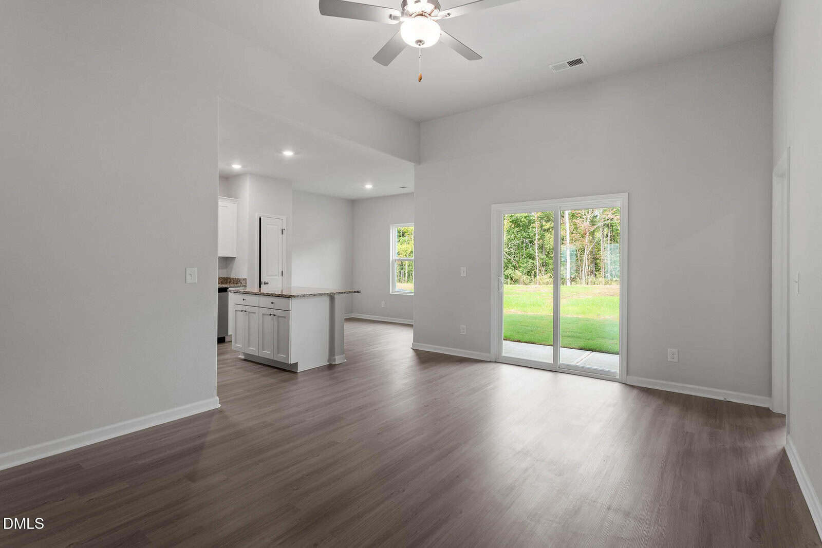Unfurnished open living area with wood flooring, ceiling fan, and sliding glass doors leading to a grassy backyard. The kitchen with white cabinets and recessed lights is visible to the left. Walls are painted light gray.