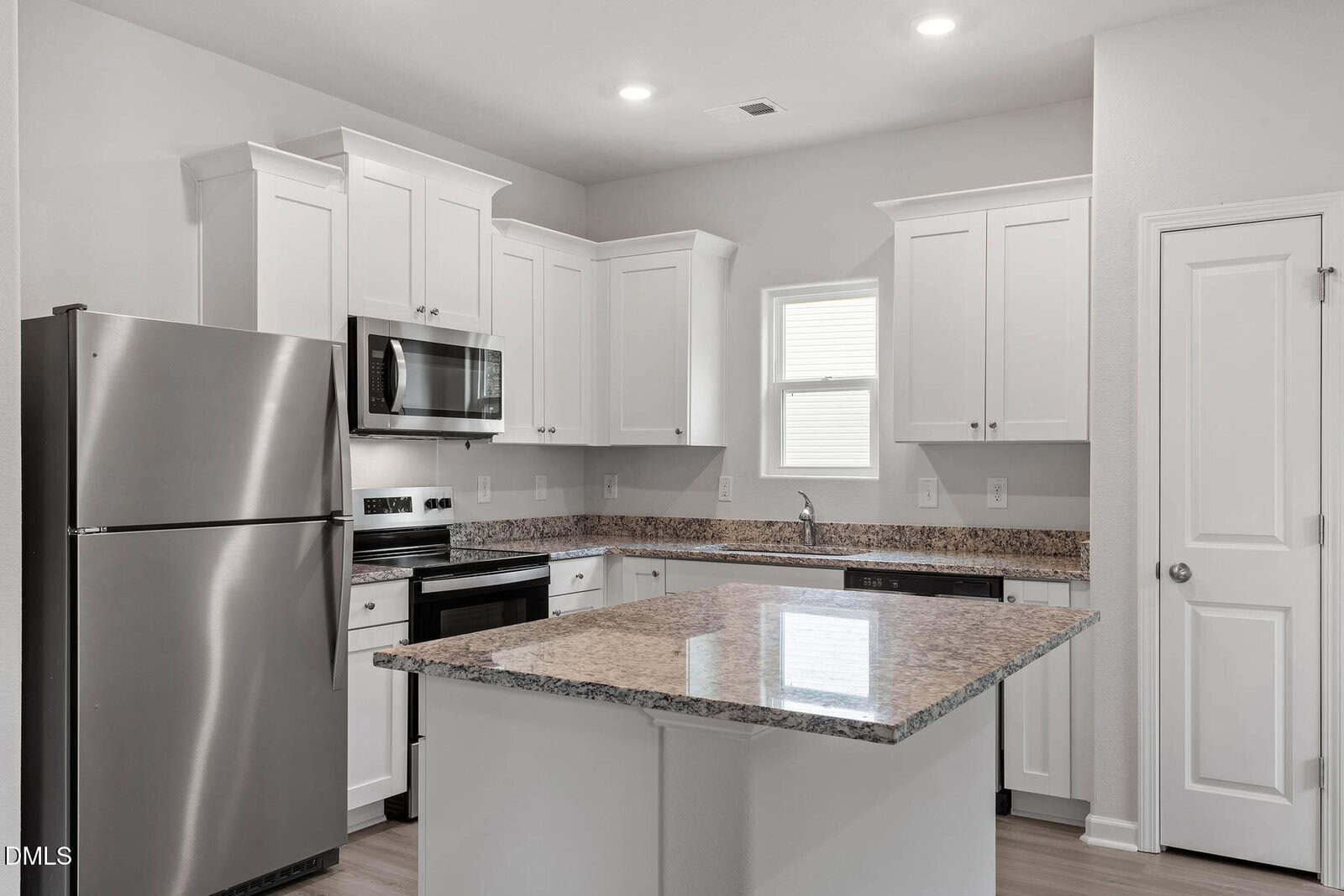 Modern kitchen with white cabinets, stainless steel appliances, granite countertops, a central island, and light wood flooring. There is a small window above the sink and a closed door to the right. The space is well-lit.