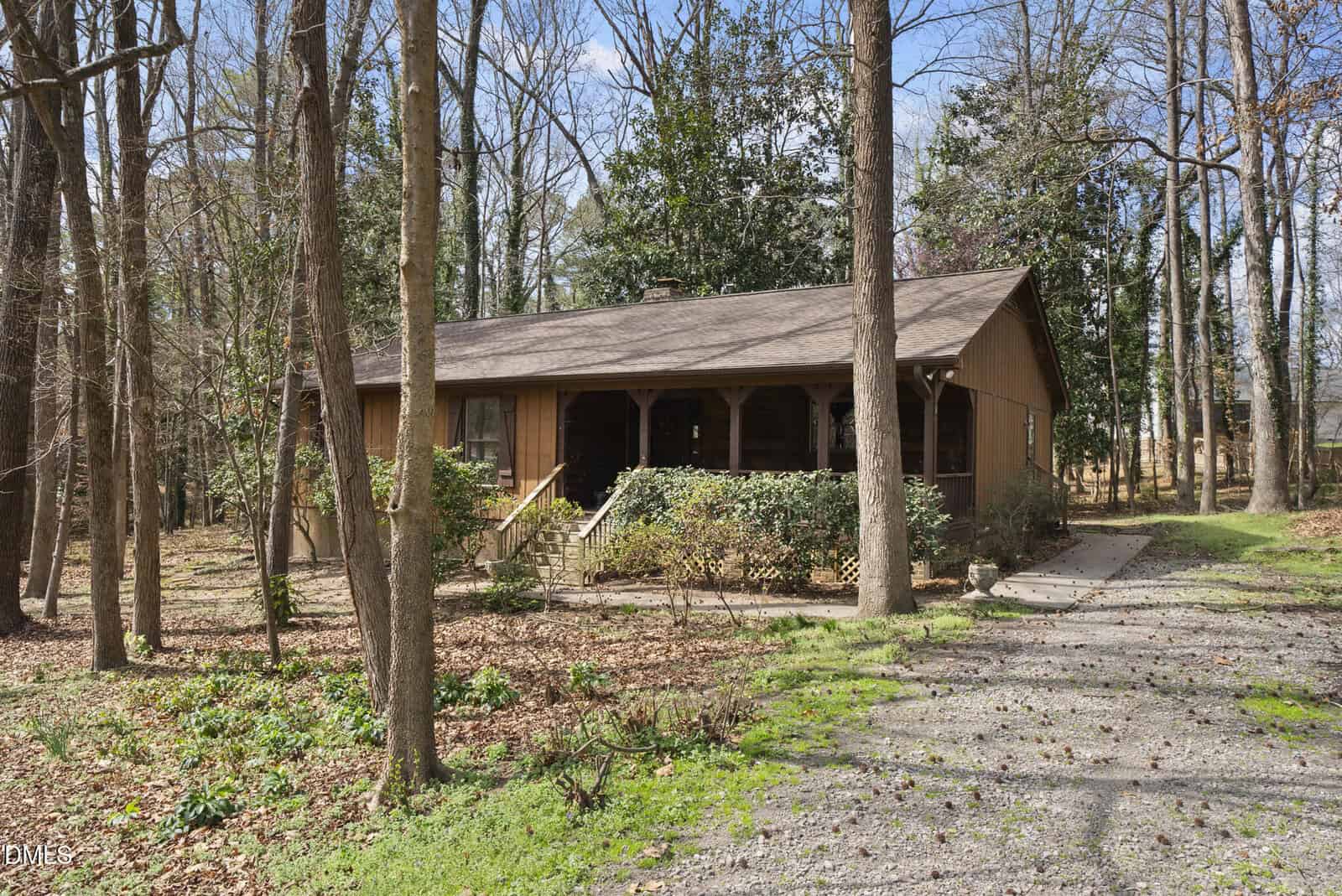 Nestled at 205 Wilder Street in Louisburg, this single-story, wood-paneled house features a charming covered front porch and is surrounded by tall trees and bushes. A gravel driveway leads up to the home, while dry leaves blanket the ground, creating a peaceful, rustic setting.
