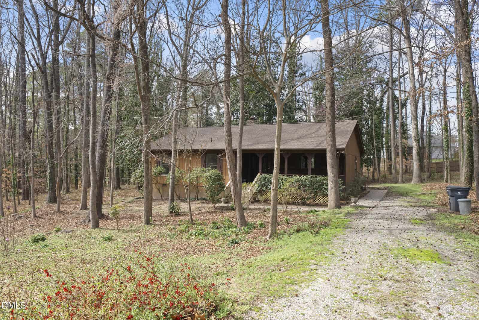 A one-story brown house with a covered front porch sits among tall leafless trees. A gravel driveway leads to the house, and there are two trash bins on the right side. The ground is covered with dry leaves and grass.