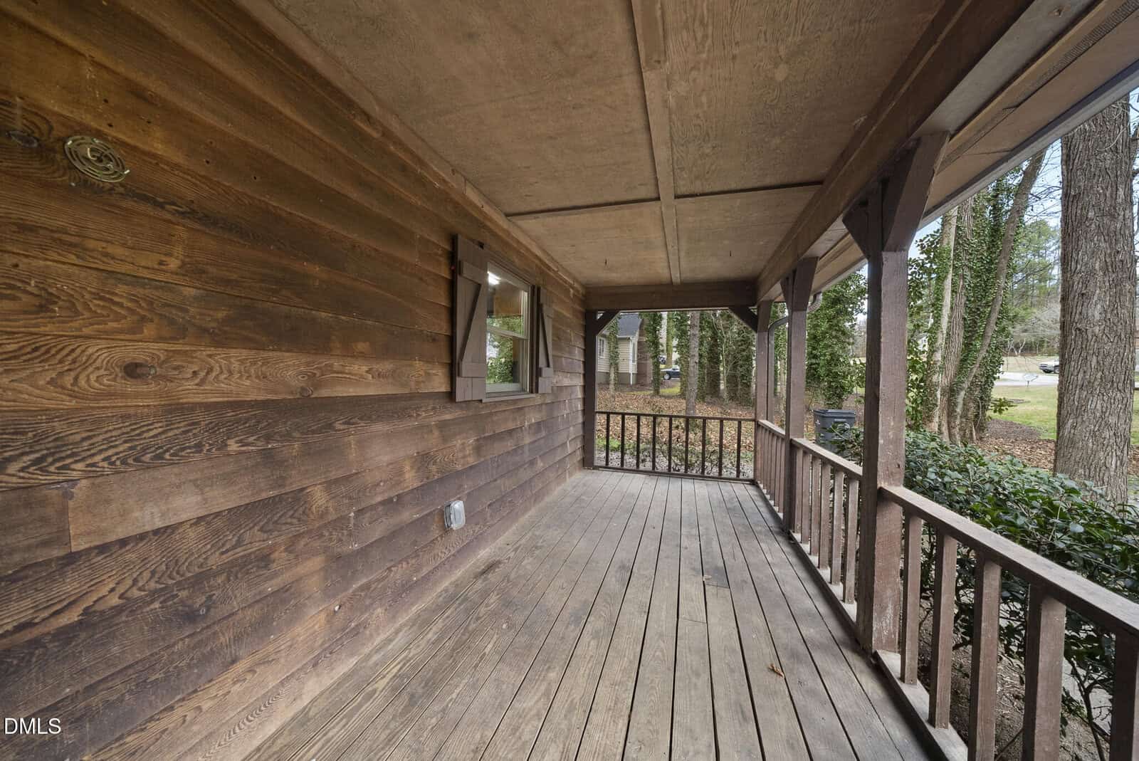 A wooden front porch with a railing, covered by a roof. The porch has weathered floorboards and overlooks a yard with trees and bushes. A small window with shutters is visible on the left wall.