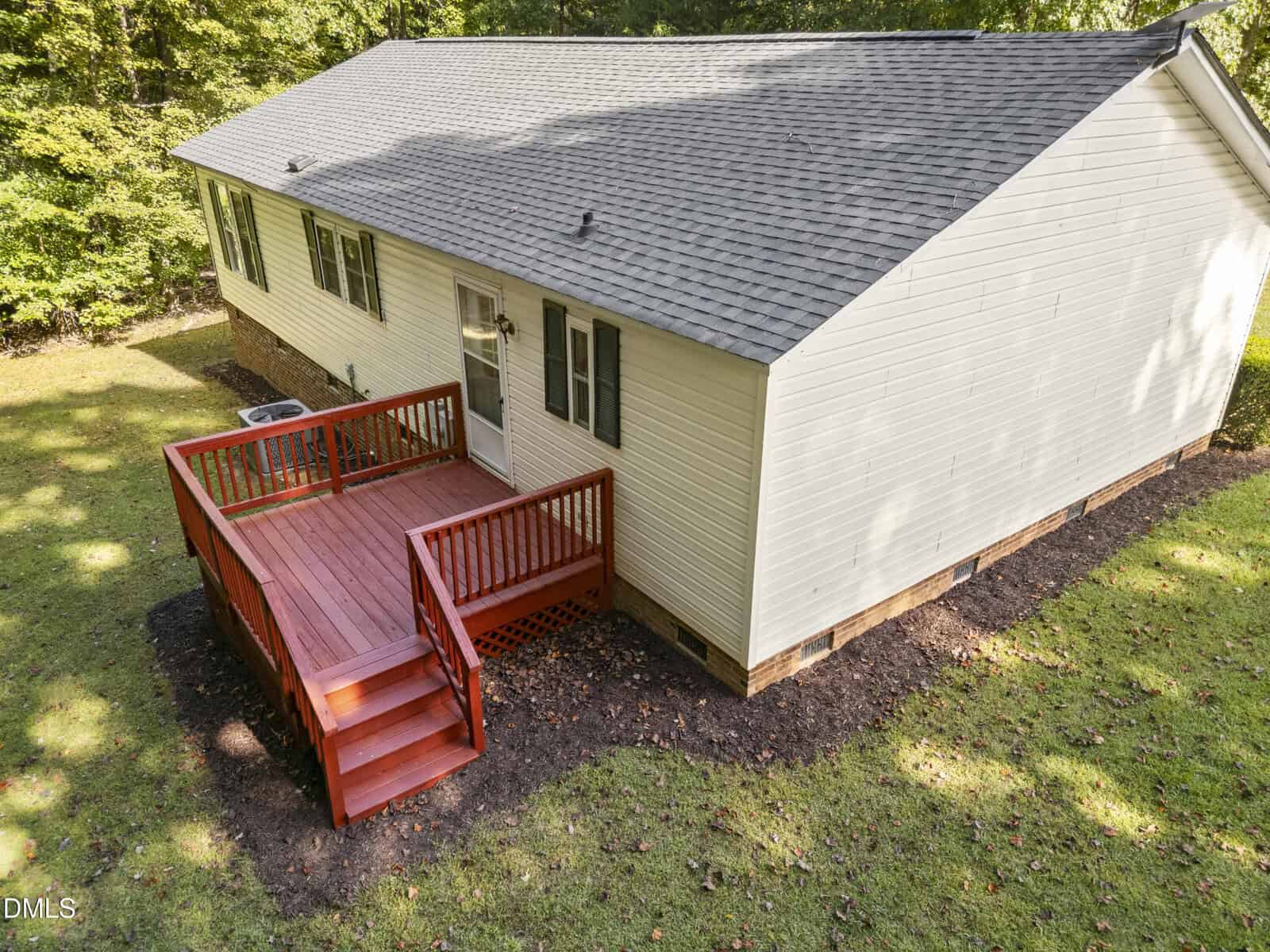 A small white house with a gray shingle roof features a red wooden deck and steps, surrounded by grass and trees in the background.