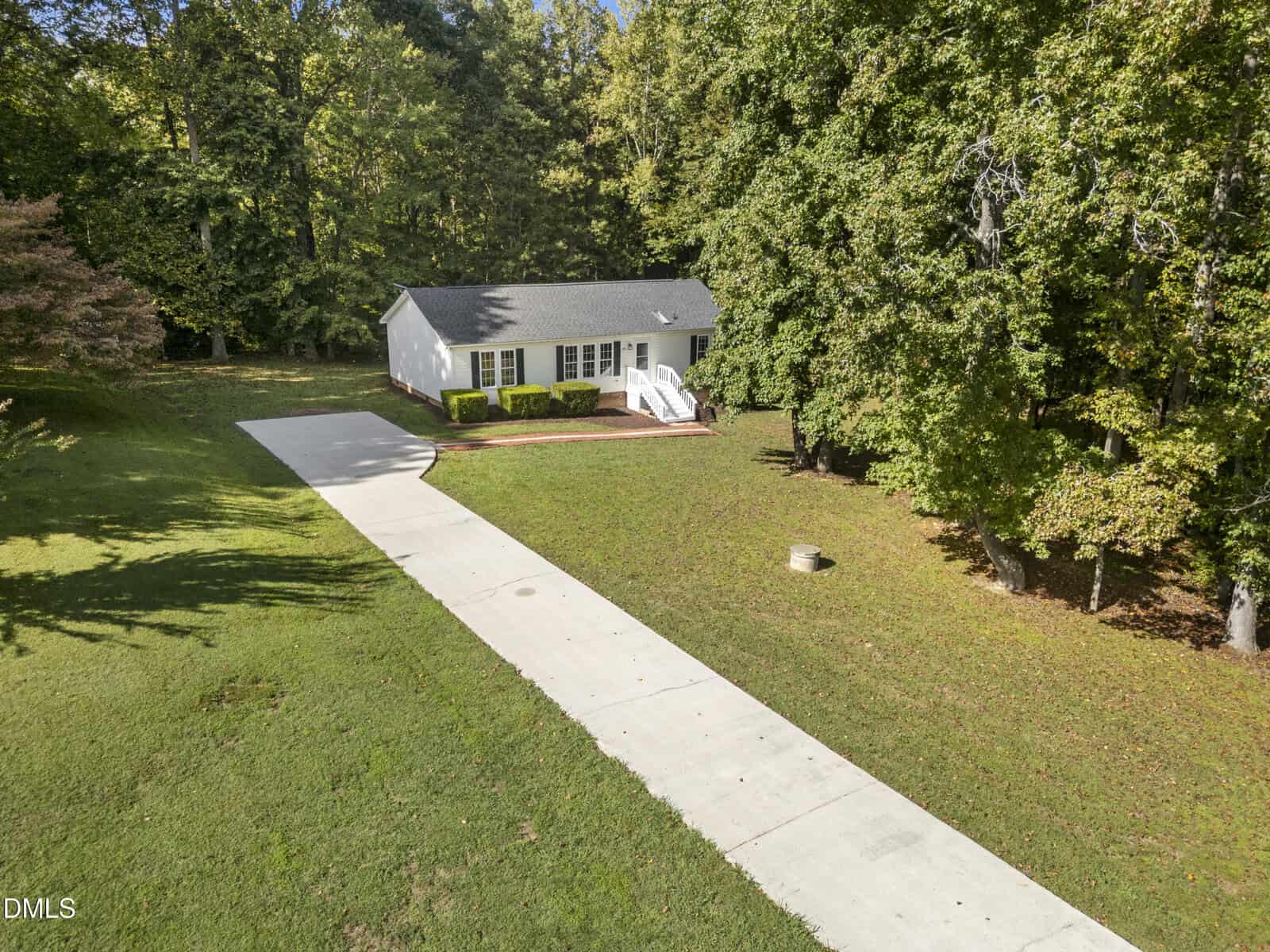 A small white house with a dark roof sits at the end of a long concrete driveway, surrounded by green grass and large trees, with a wooded area in the background.
