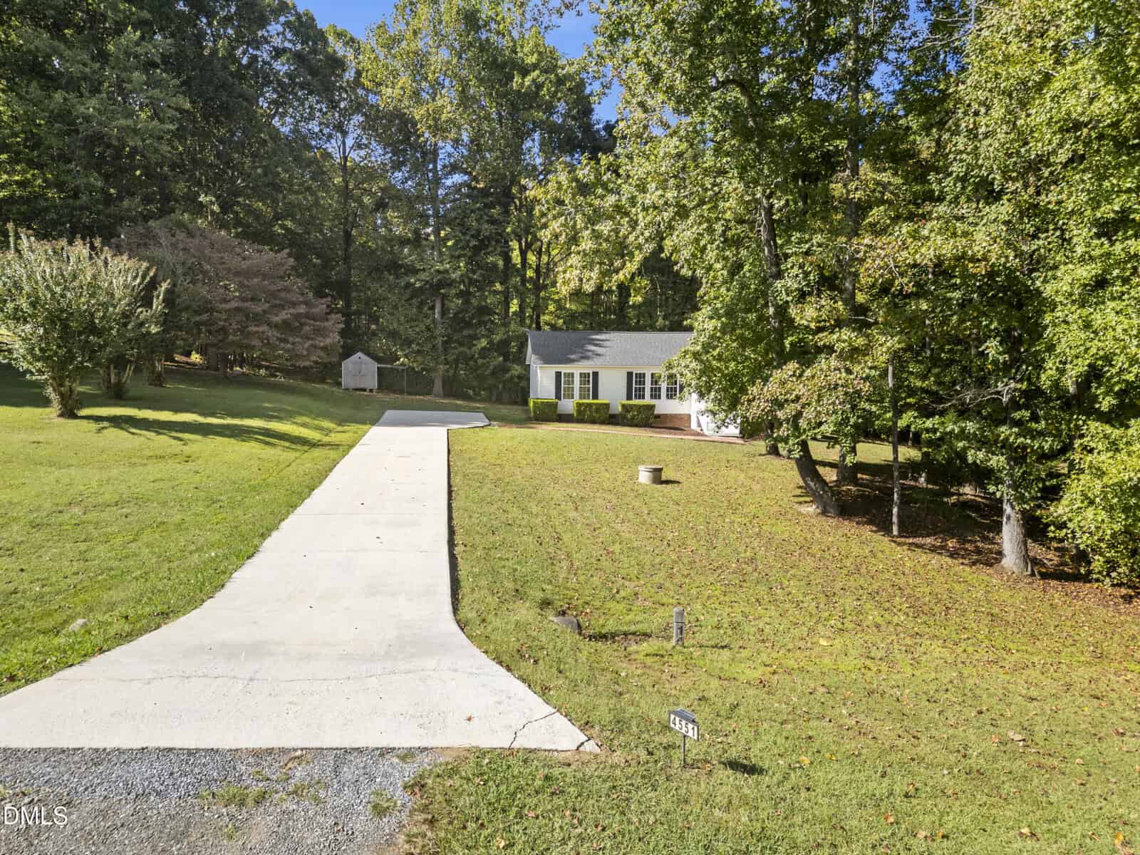 A single-story white house sits at the end of a long concrete driveway, surrounded by a lawn and trees. A small shed is visible in the background, and the sky is clear and sunny.