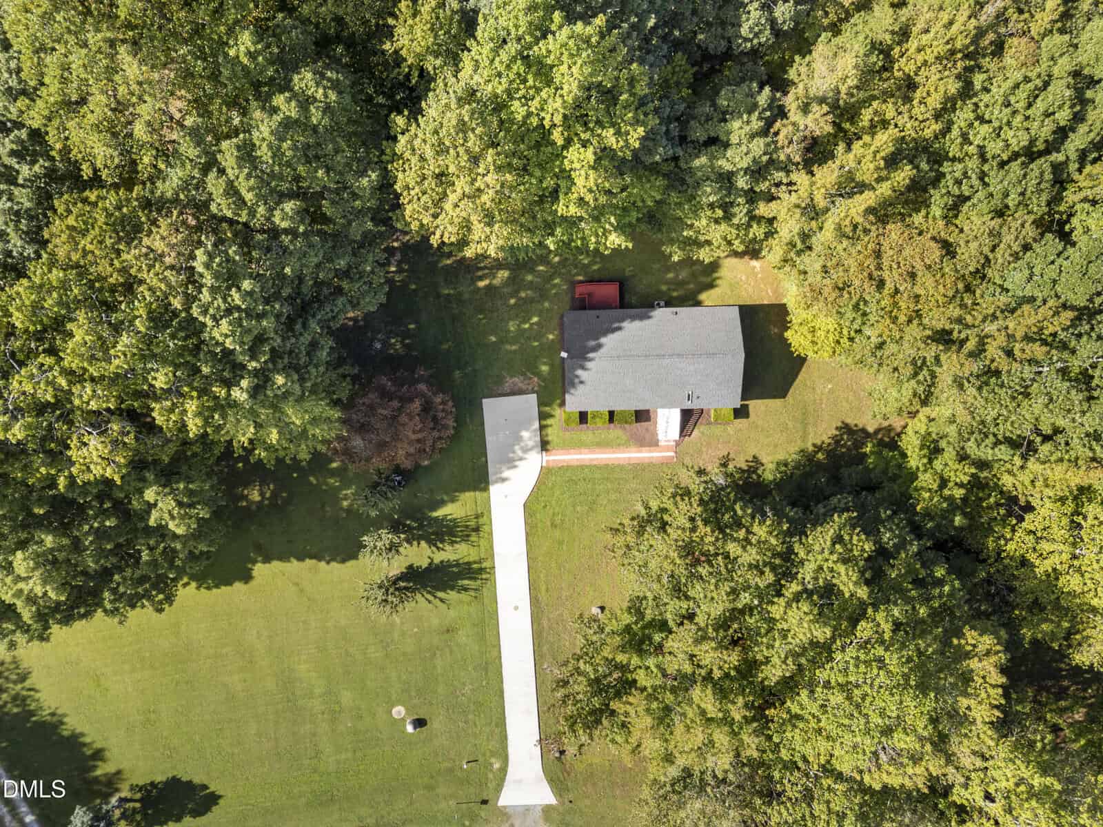 Aerial view of a single-story house with a gray roof and a long white driveway, surrounded by green grass and dense trees on all sides. The property appears well-maintained and shaded by mature trees.