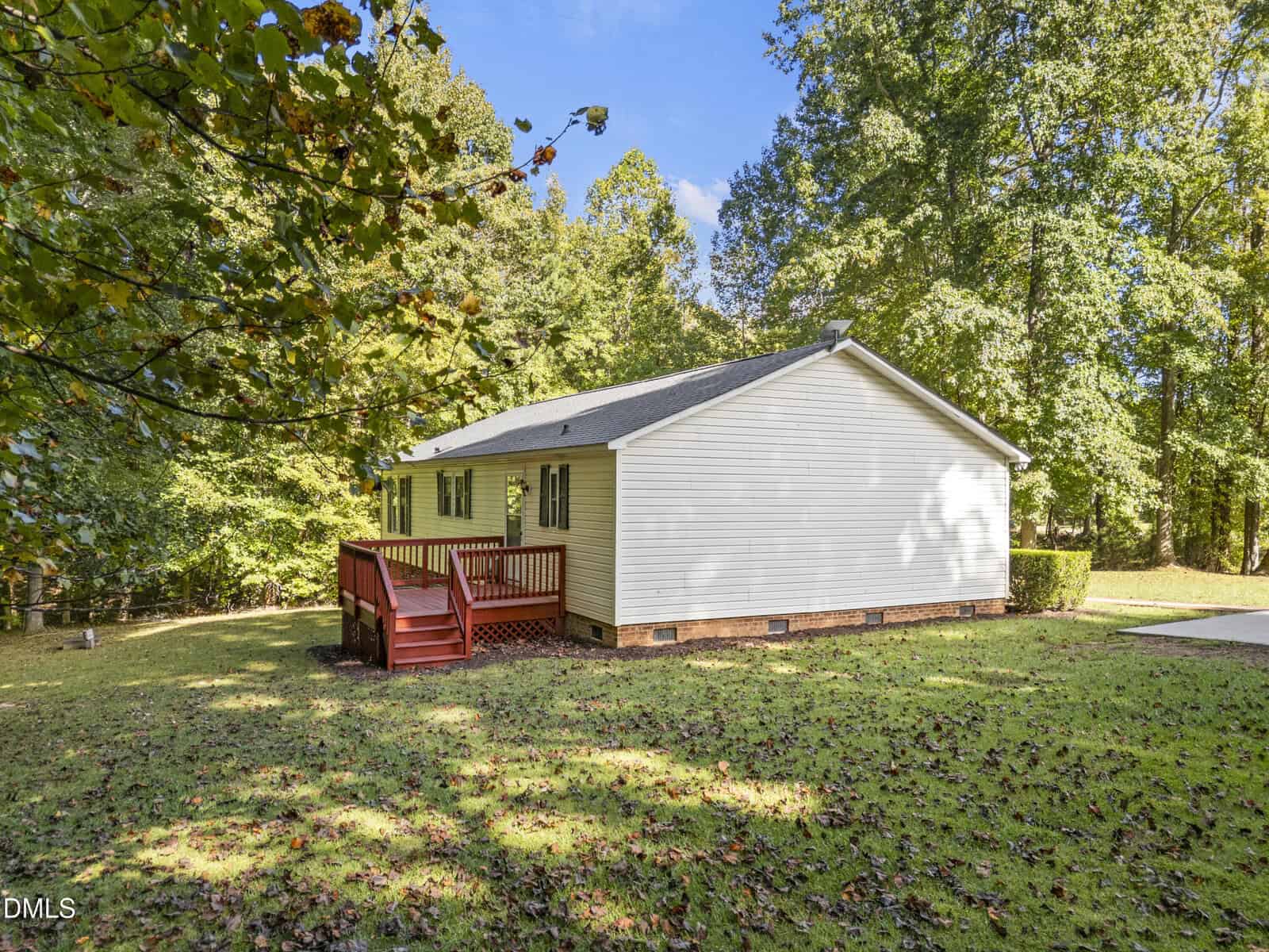 A small white house with a gray roof and red wooden deck sits on a grassy lawn, surrounded by tall green trees under a bright blue sky.