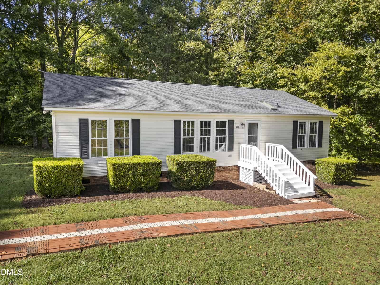 A small white single-story house with black shutters, gray roof, and front steps leading to a white porch. The yard features trimmed hedges, a red brick pathway, and is bordered by green grass and trees.
