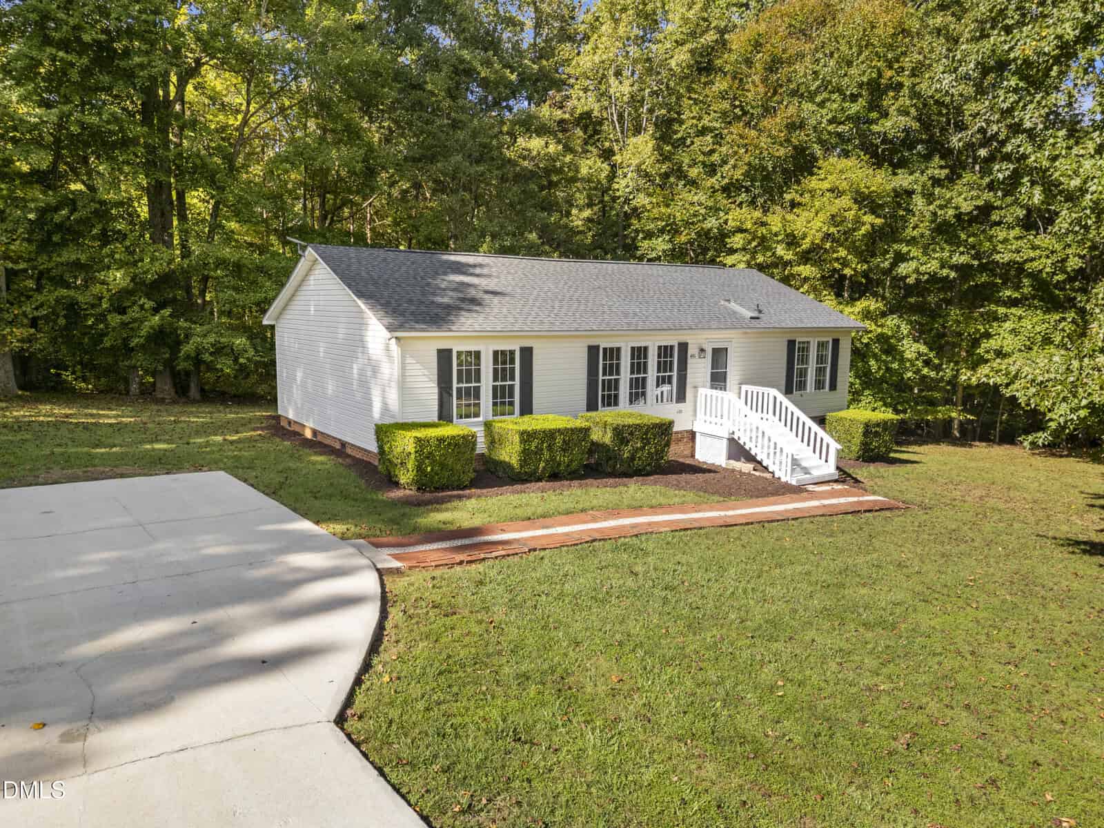 A small white single-story house with gray shingles on 4551 Sugar Maple Road, Oxford, featuring white steps, trimmed bushes, a concrete driveway, and a grassy lawn edged by trees in the background.