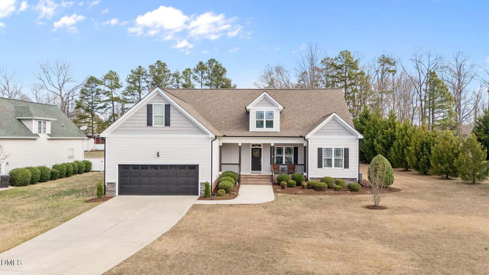 Located at 10 Tall Cedar Lane in Youngsville, this two-story suburban house features a gray exterior, black garage door, and charming front porch. Neatly trimmed shrubs line the spacious yard, which boasts lush grass and a few small trees. A concrete driveway leads to the garage, while the property is bordered by tranquil woods in the background.