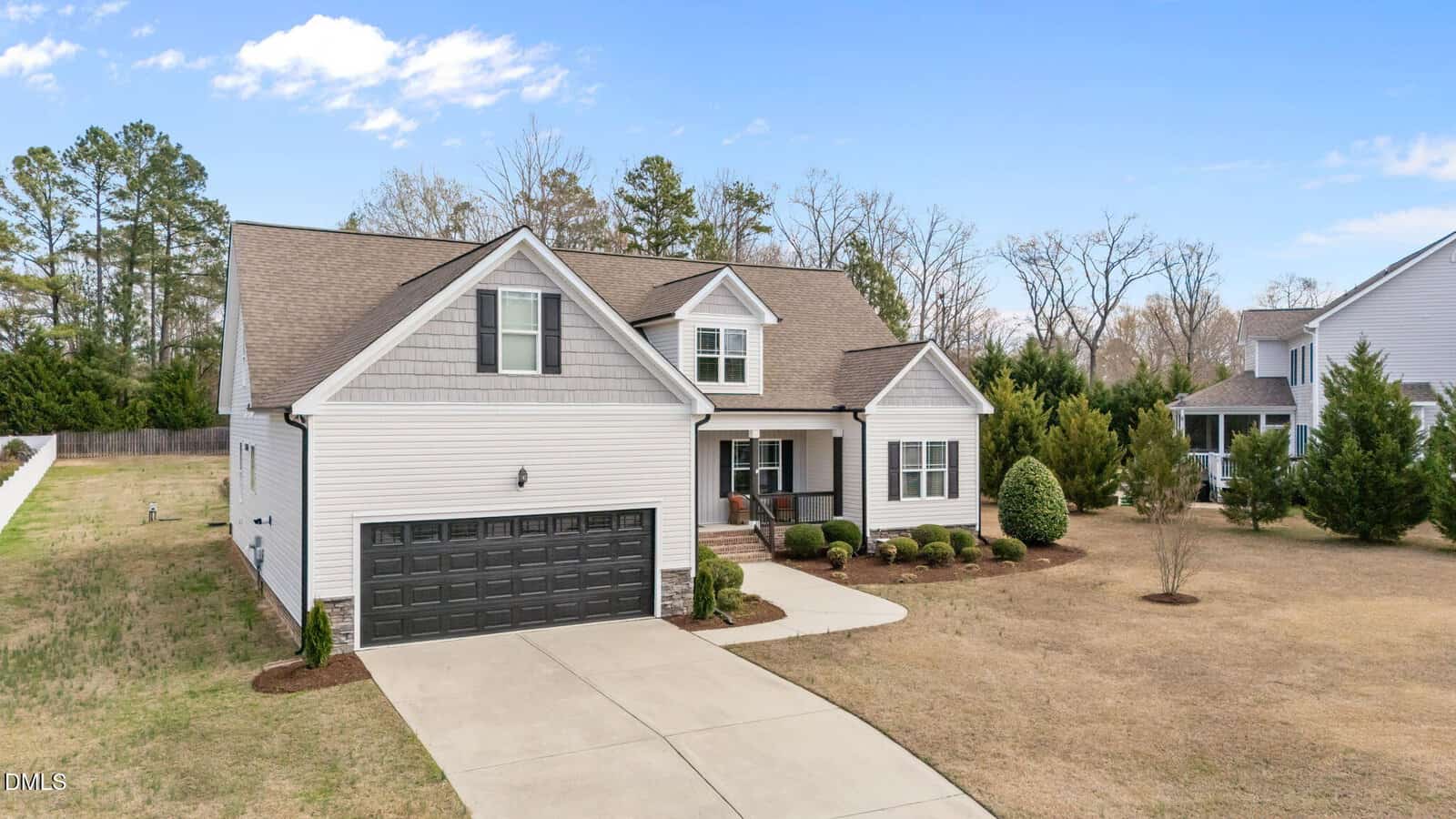 A two-story suburban house with gray siding, black garage door, front porch, and a driveway. The yard is mostly grass with minimal landscaping, and trees are visible in the background.