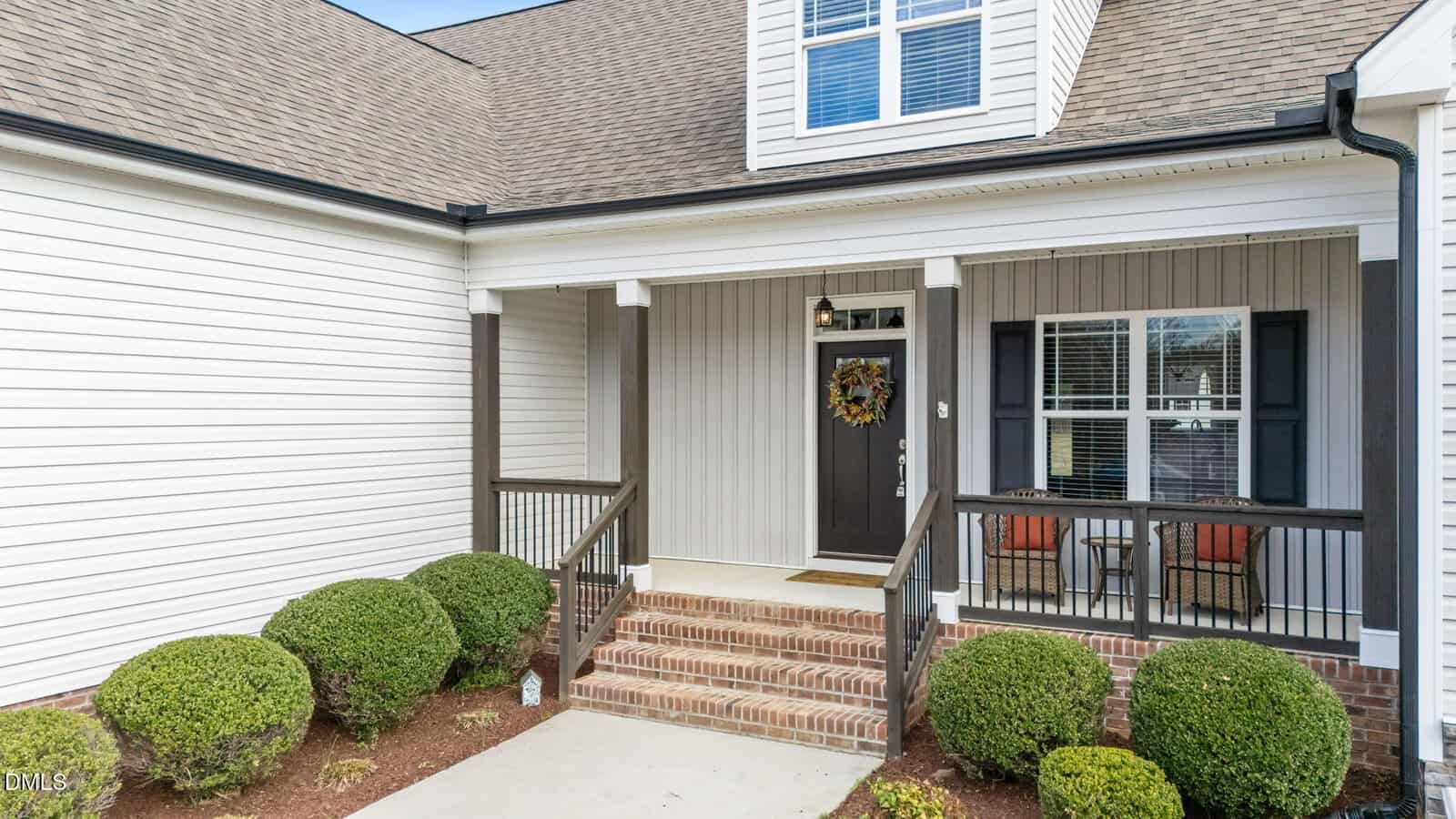 A front porch with brick steps, white columns, and a black door with a wreath. Two chairs and a small table are on the porch. Shrubs line the walkway. The house has beige siding and a gabled roof.