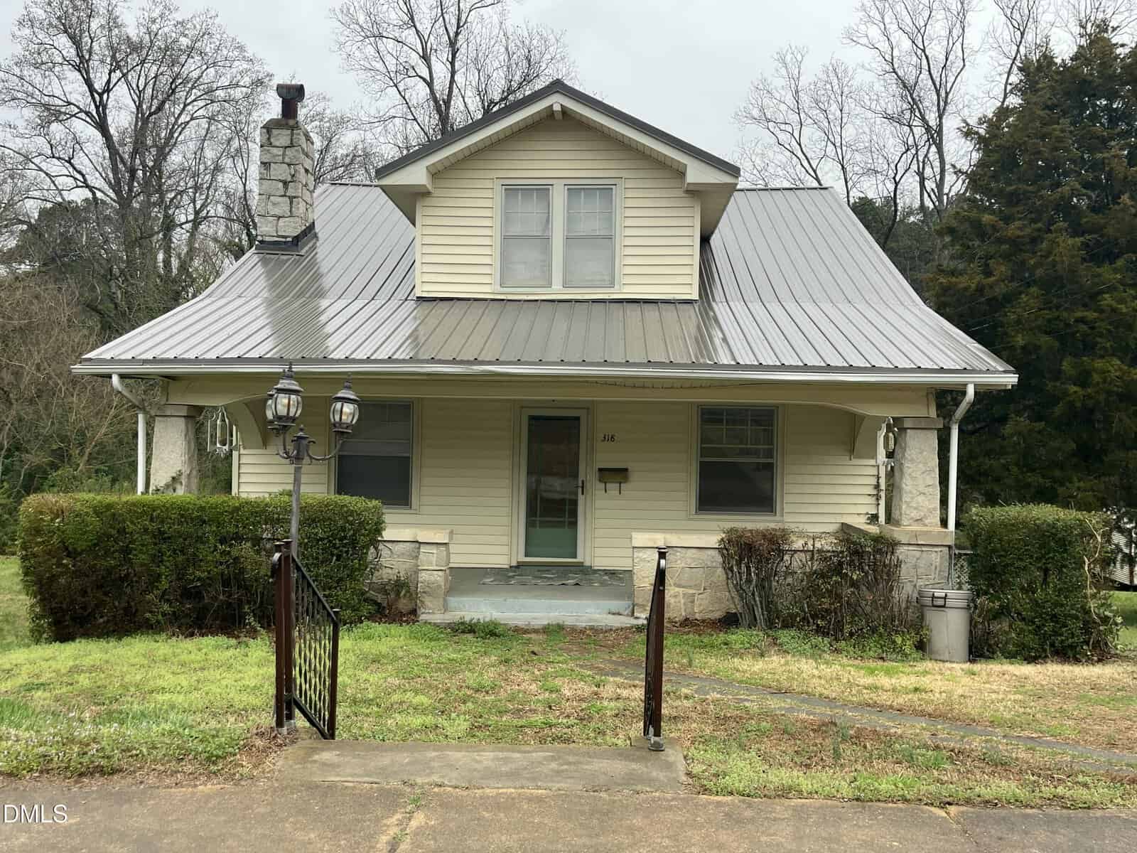 A beige house with a metal roof sits at 318 W Franklin Street in Warrenton, featuring two upper windows, a small porch, and shrubs in front. Two lamp posts stand near the entrance, while bare trees frame the background on a cloudy day.
