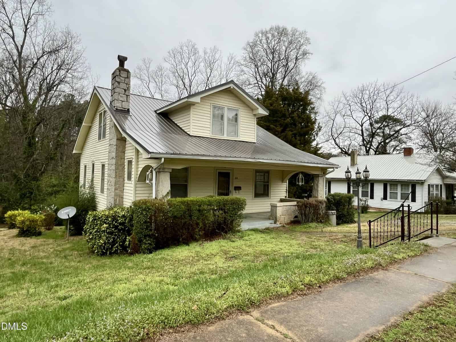A beige two-story house with a metal roof, a stone chimney, and a covered front porch, surrounded by grass, bushes, a driveway, and a black gate. Other houses and bare trees are visible in the background.