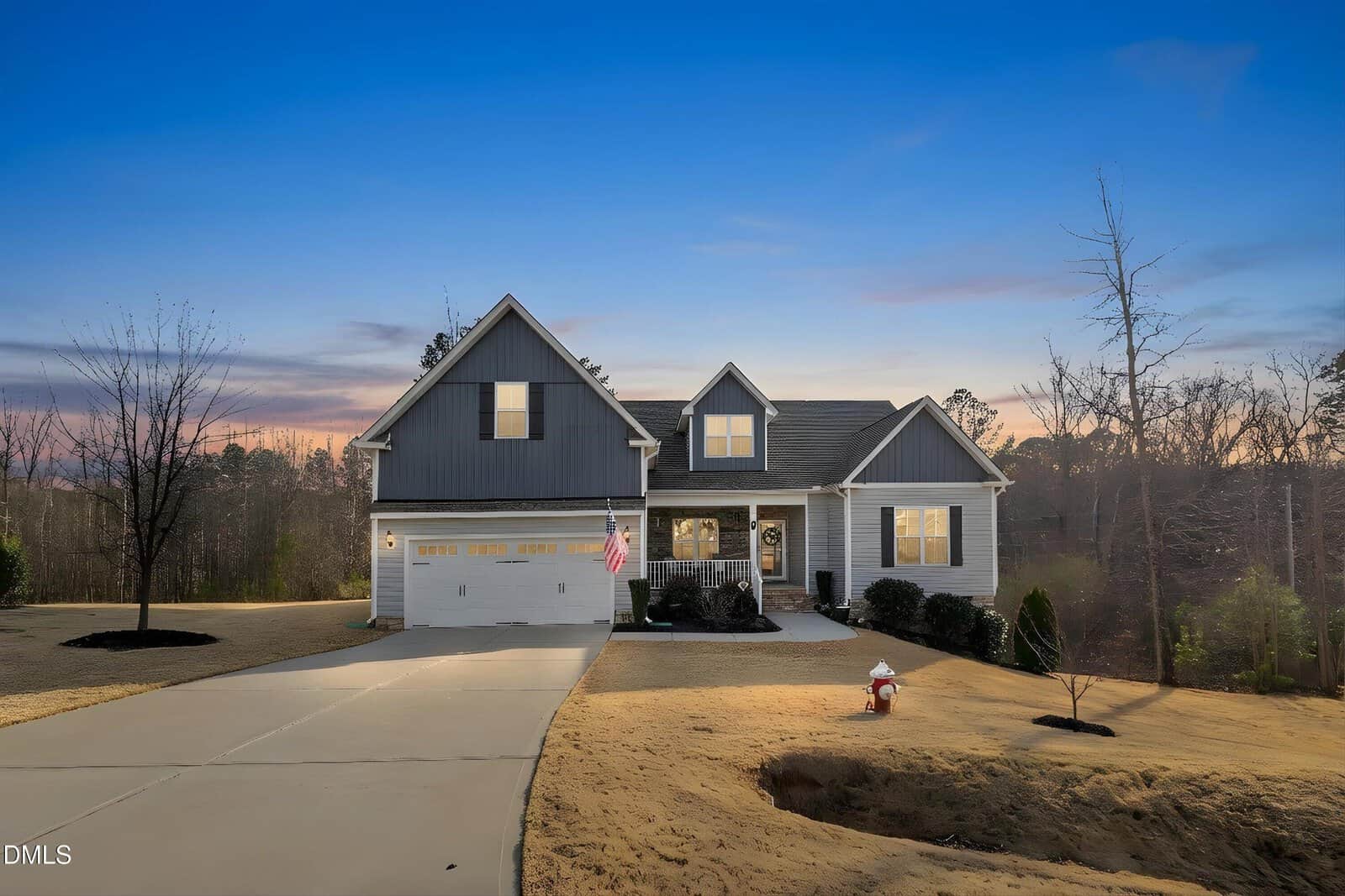 Two-story suburban house with a gray and white exterior, double garage, and front porch, set on a large lot with minimal landscaping and surrounded by trees at sunset. An American flag hangs near the front door.