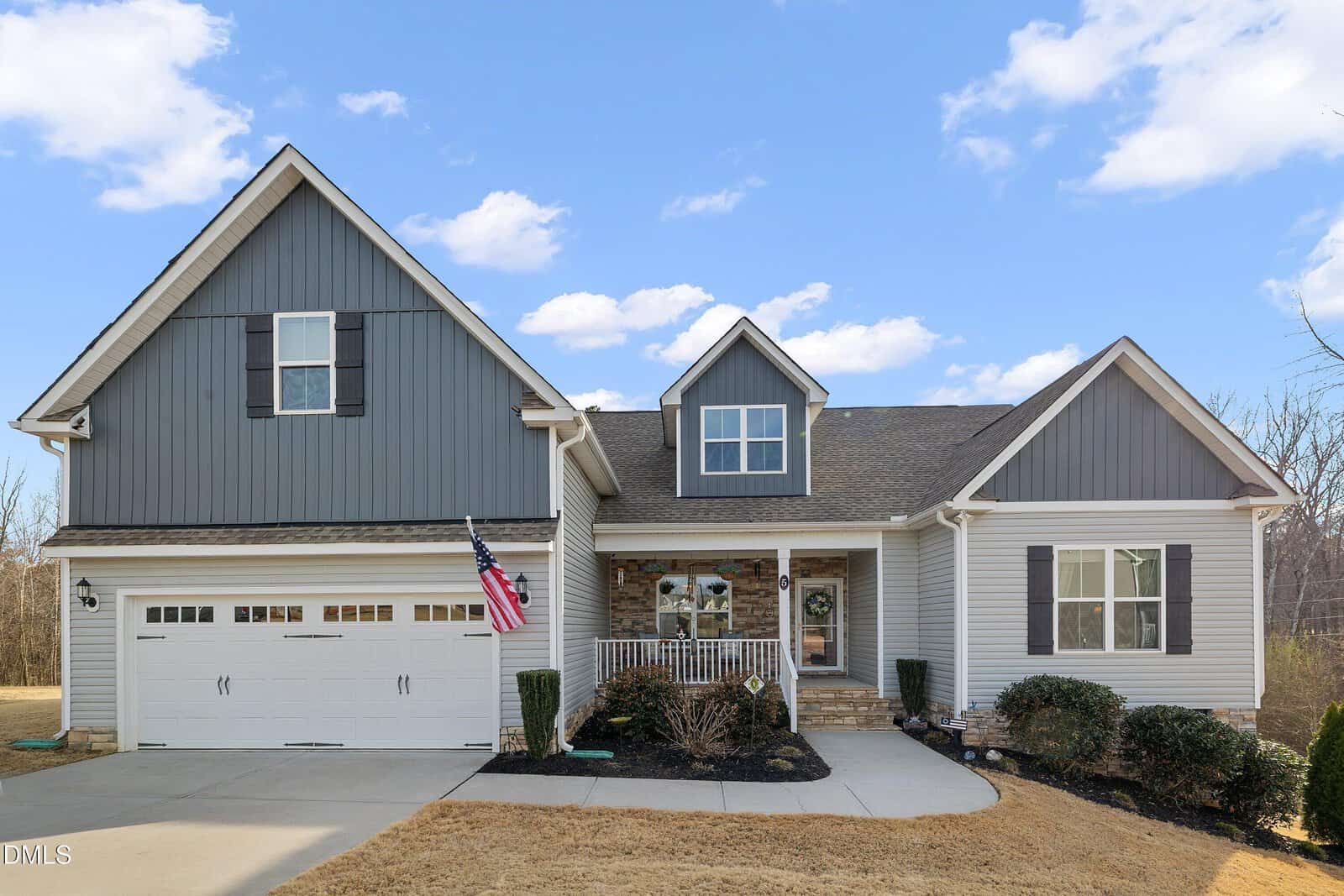A modern, two-story suburban house with gray siding, stone accents, a covered front porch, and an American flag by the white garage. The yard has a driveway, trimmed bushes, and a blue sky with clouds in the background.