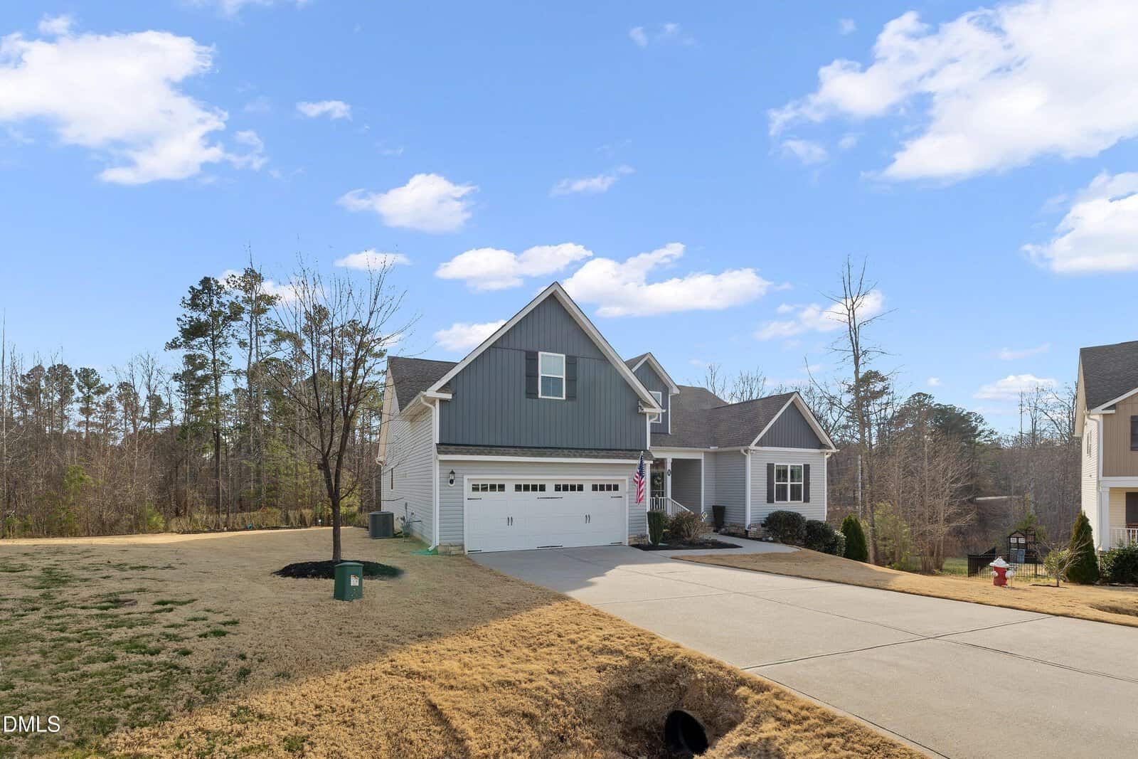 A modern gray house with a two-car garage, white trim, and a sloped driveway sits in a suburban neighborhood with trees and a clear blue sky in the background.