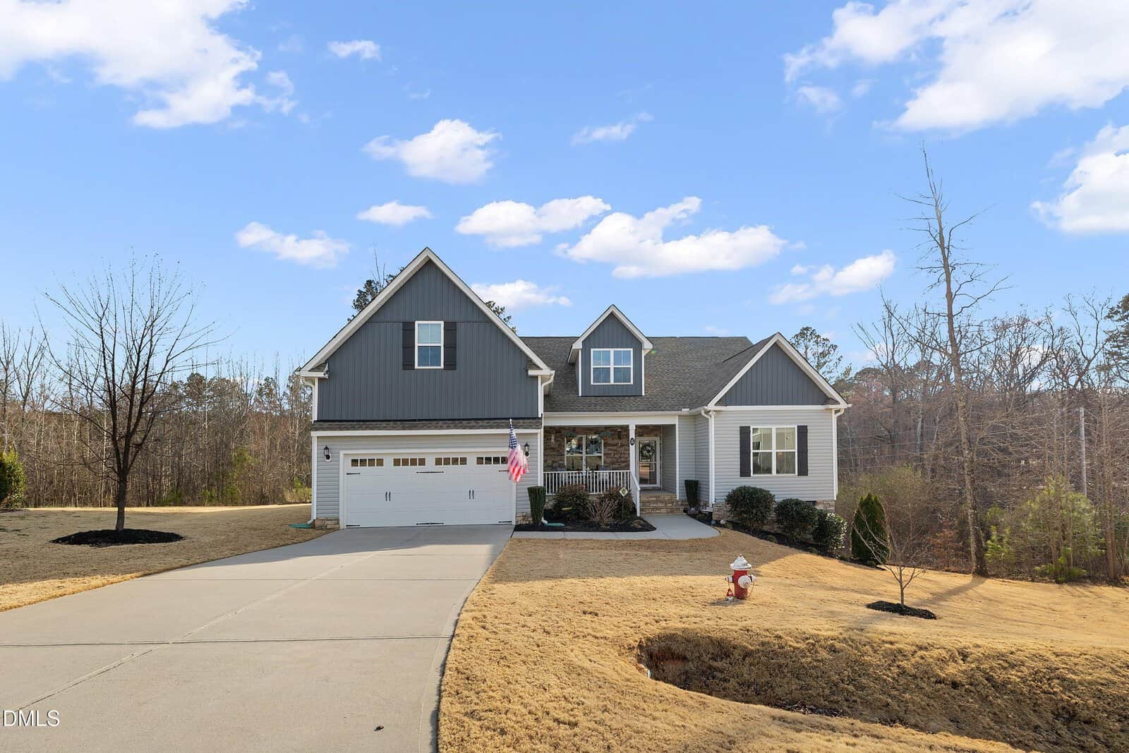 A modern gray and white two-story suburban house with a double garage, American flag, and wreaths on the door, sits on a manicured lawn with a driveway and a red fire hydrant, surrounded by trees under a blue sky.
