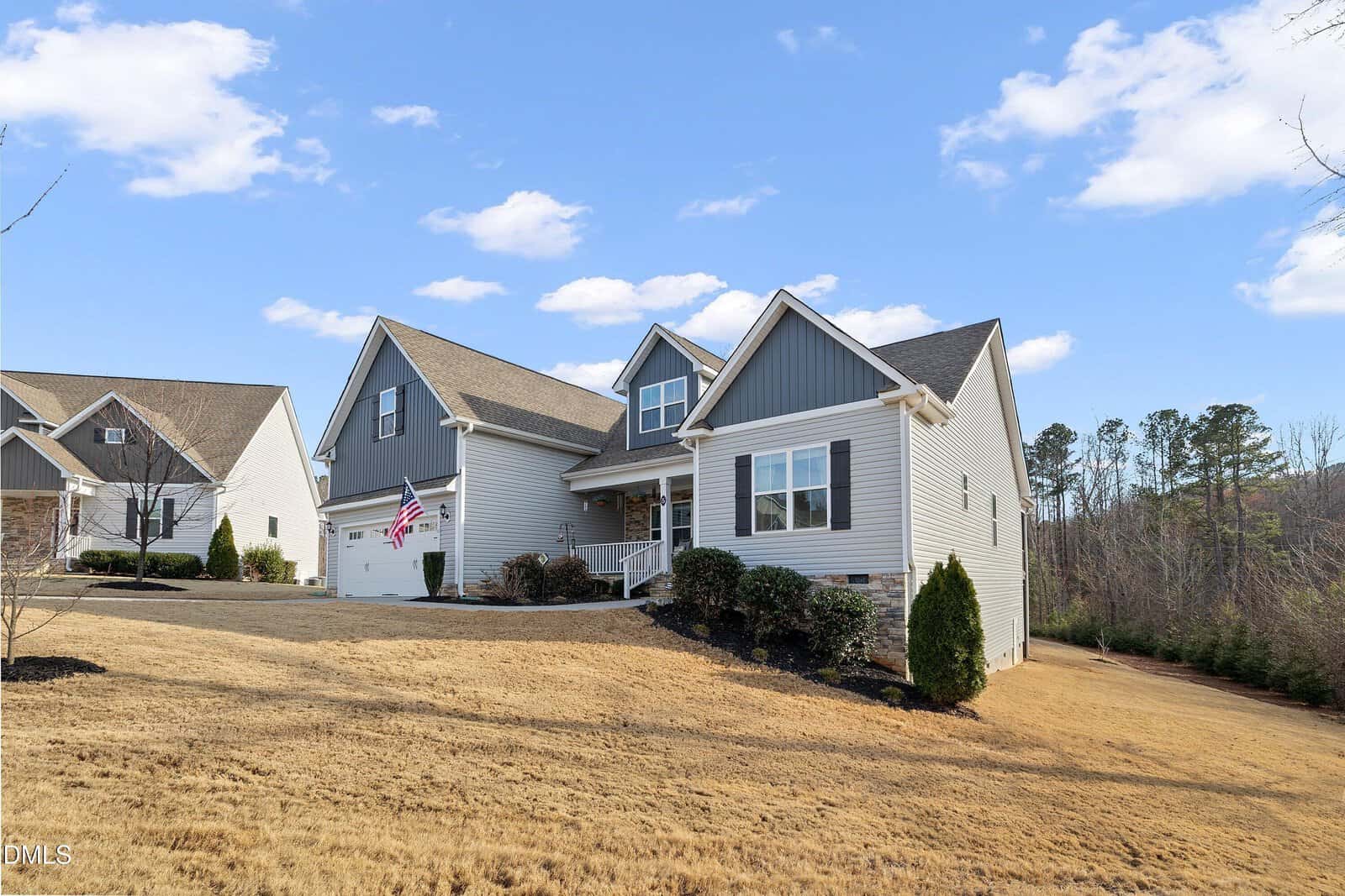 A light gray, two-story suburban house with white trim and a double garage stands on a sloped, yellow-brown lawn. An American flag hangs near the garage. The sky is partly cloudy with scattered trees in the background.