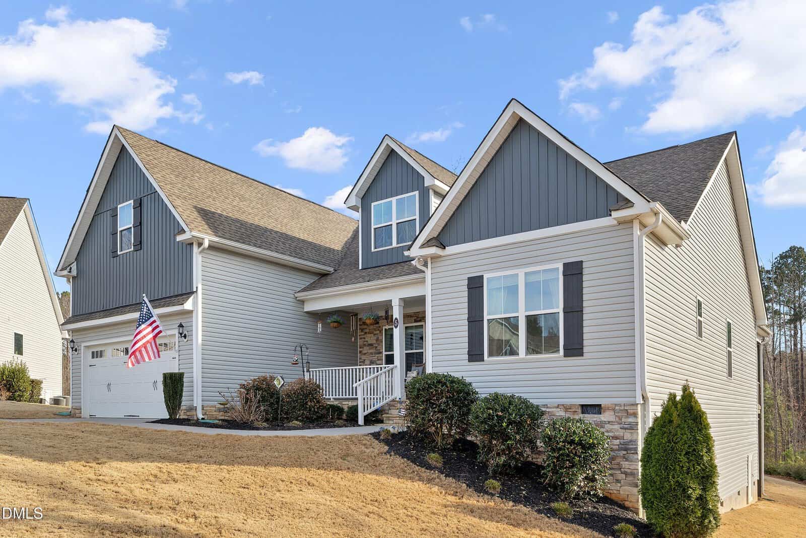 A two-story suburban house with gray siding, white trim, a covered front porch, black shutters, and a front lawn. An American flag hangs near the entrance, and shrubs line the front of the house.