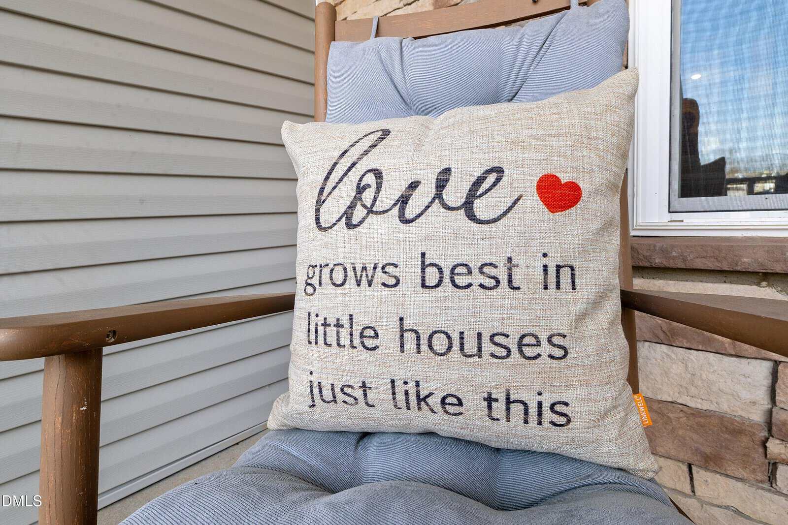 A beige pillow with text reading “love grows best in little houses just like this” and a red heart sits on a cushioned wooden chair on a porch near siding and a window.