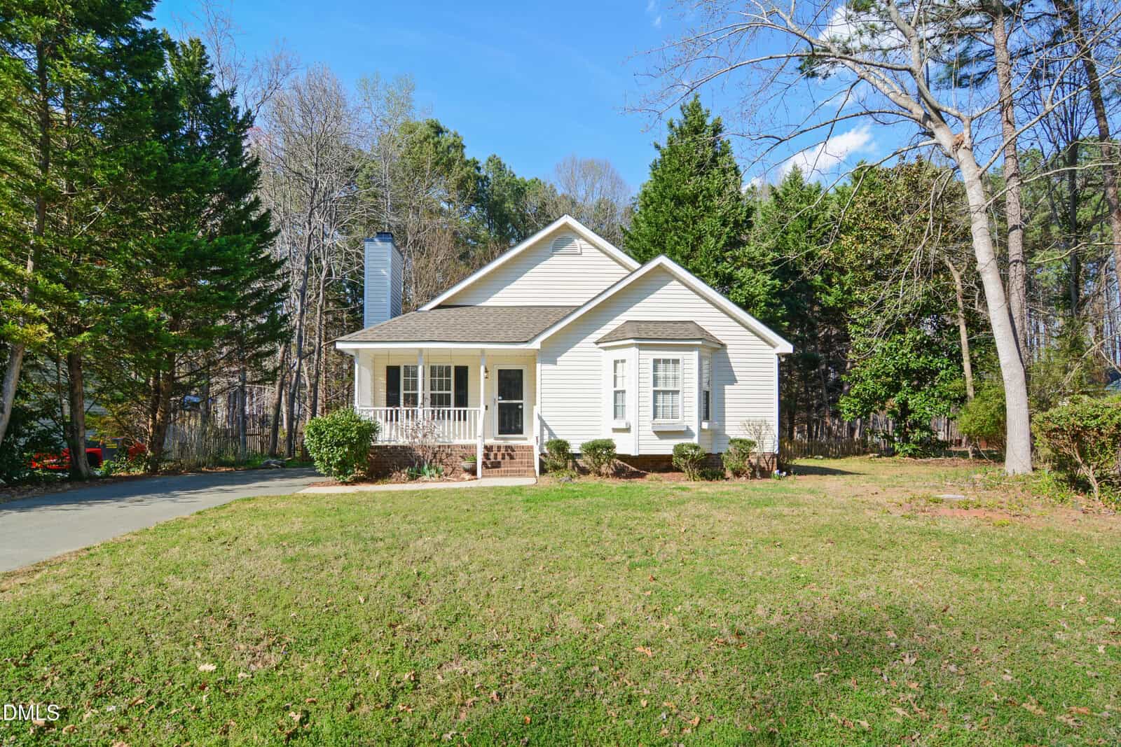 Single-story house with beige siding and a covered front porch, surrounded by grass and tall trees. A curved driveway leads to the left of the house under a clear blue sky.