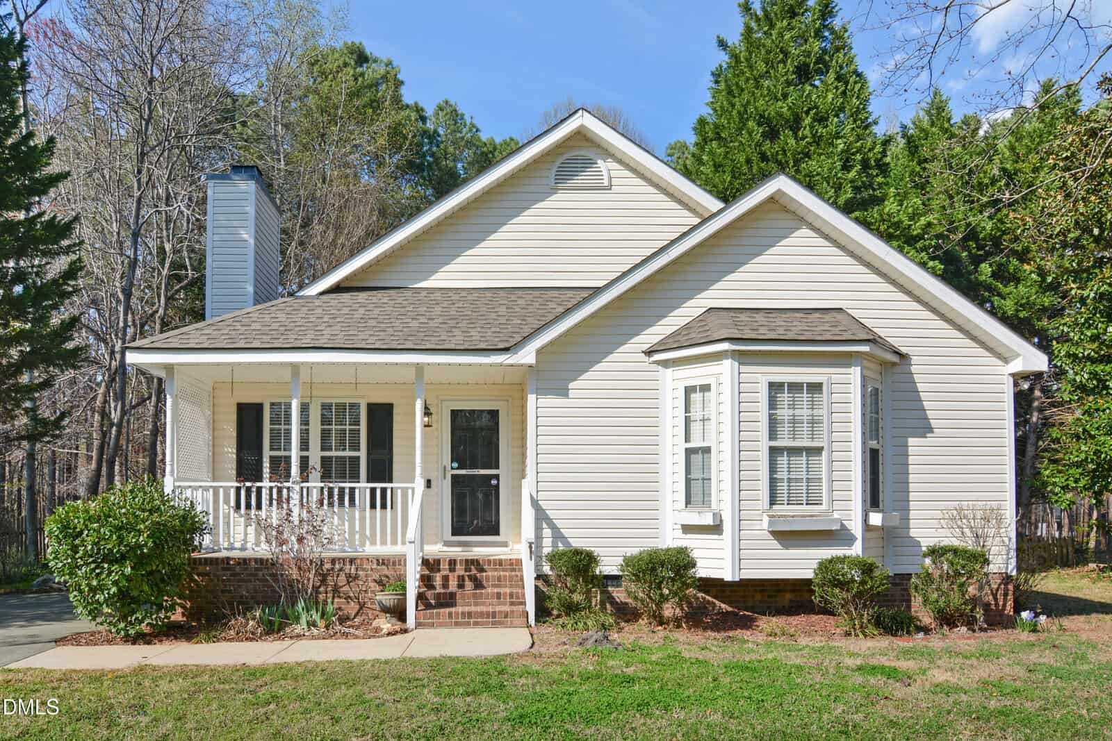 Charming single-story beige house at 110 Eagle Stone Ridge, Youngsville, featuring a covered front porch, brick steps, black front door, bay window, and crisp white trim. The yard is adorned with shrubs and bordered by trees beneath a clear blue sky.