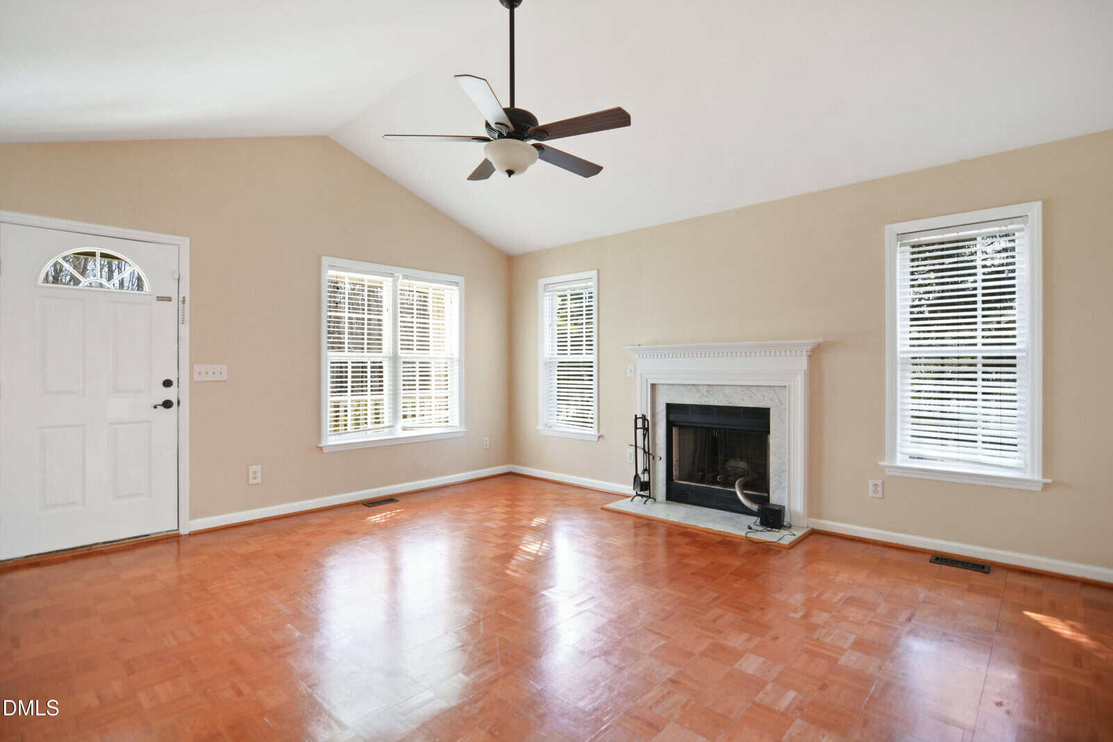 Empty living room with light beige walls, three tall windows with white blinds, a white front door, a white fireplace, wood floor, vaulted ceiling, and a black ceiling fan with lights.