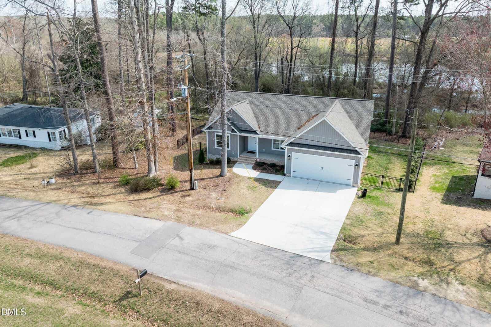A gray single-story house with a two-car garage and a wide driveway sits along a paved, tree-lined street. The yard is mostly bare with a few shrubs. Nearby houses and a wooded area are visible in the background.