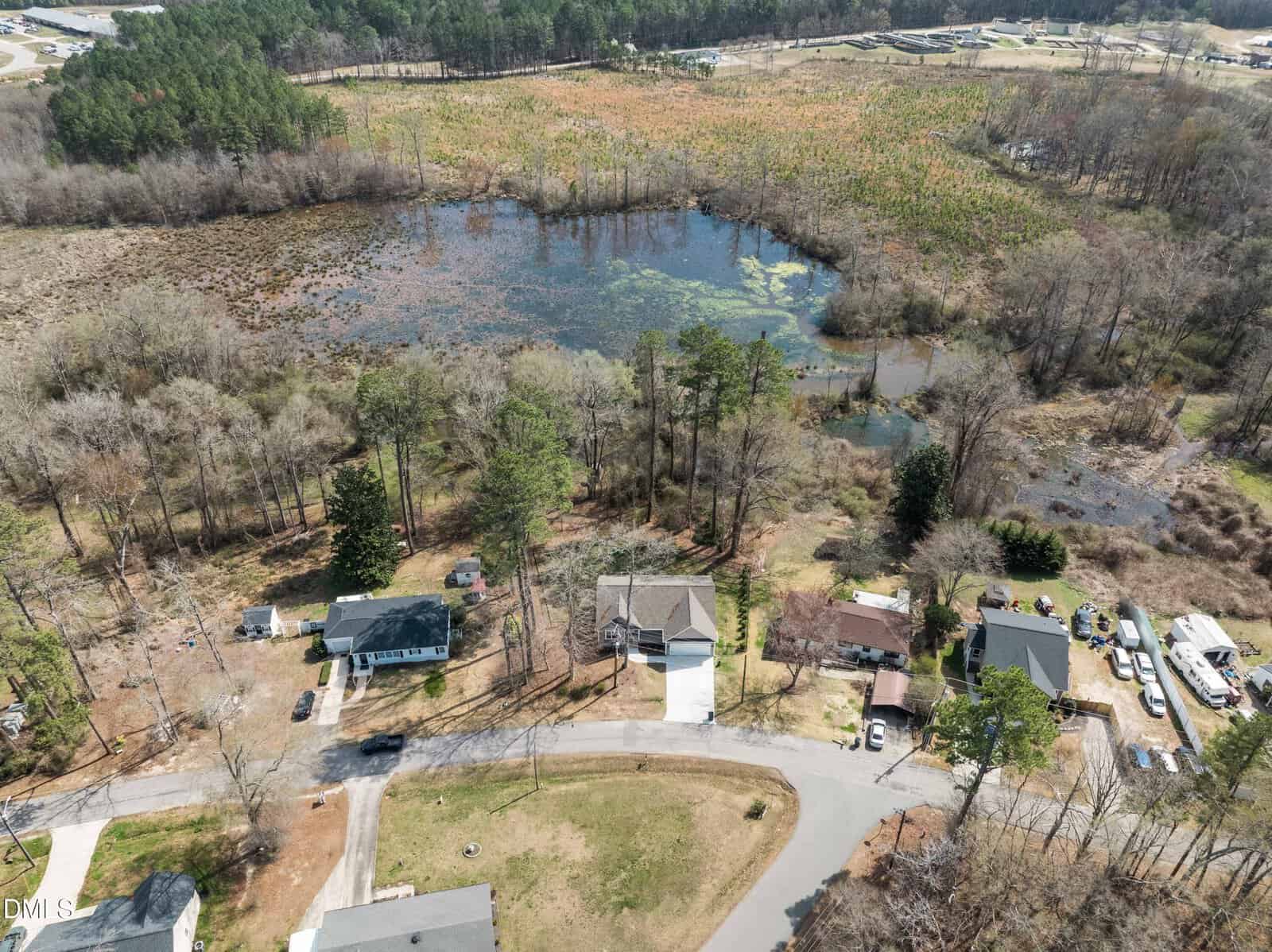 Aerial view of several houses bordering a small lake surrounded by trees and marshland, with a residential road curving in the foreground and forested areas in the background.