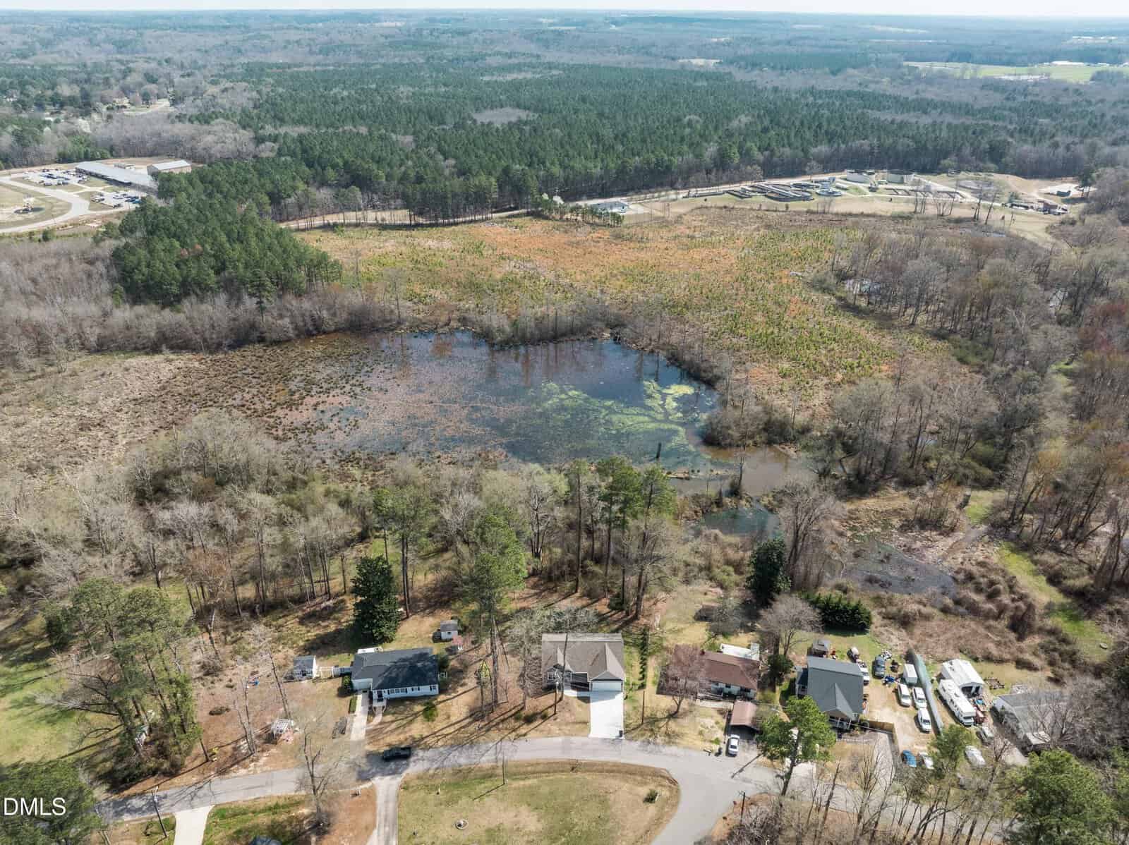 Aerial view of houses bordering a small pond surrounded by trees and vegetation, with a wooded area and open fields in the background under a partly cloudy sky.