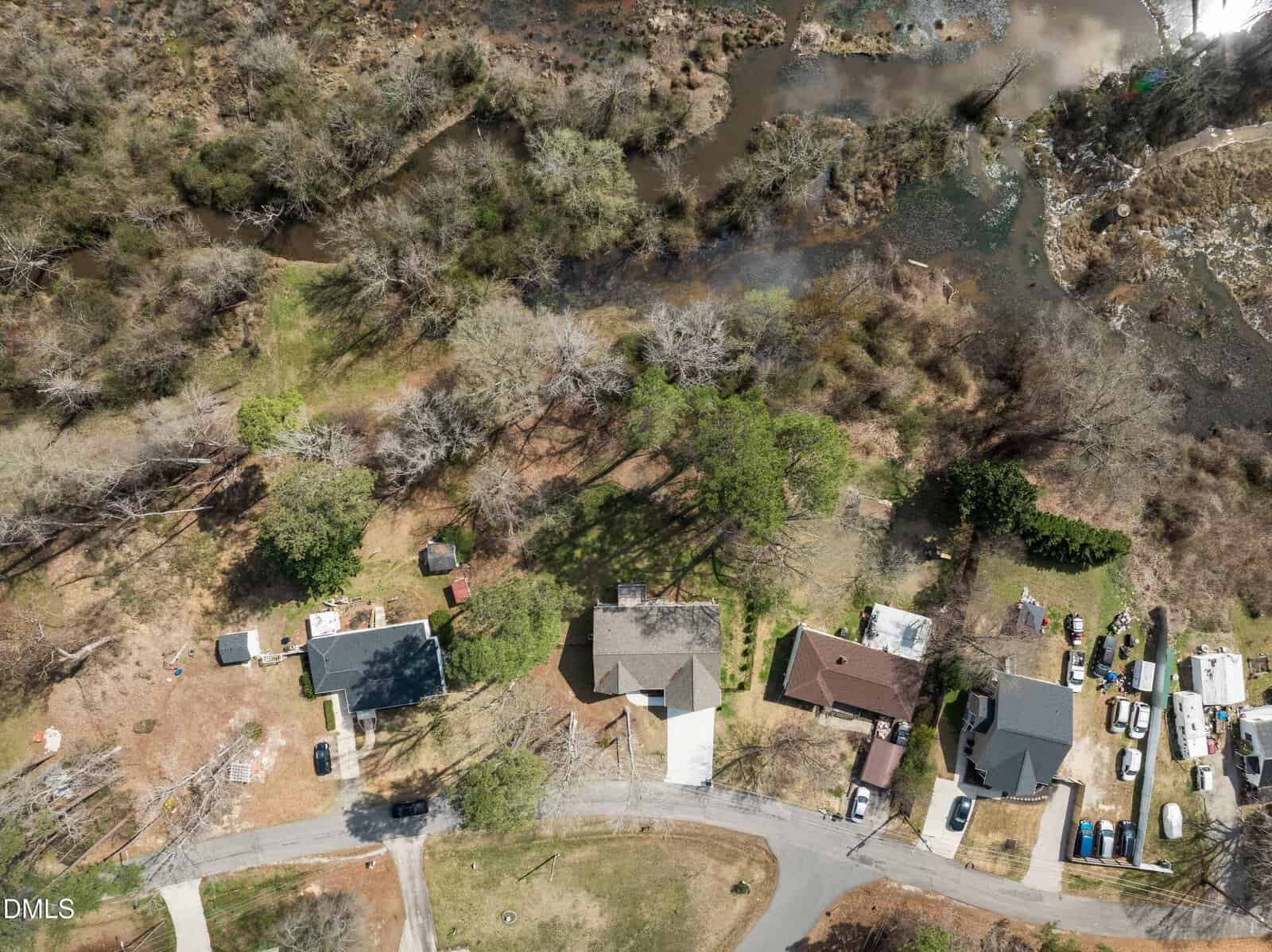 Aerial view of several houses along a curved street near a wooded area and a pond, with yards, vehicles, and scattered trees visible in the neighborhood.