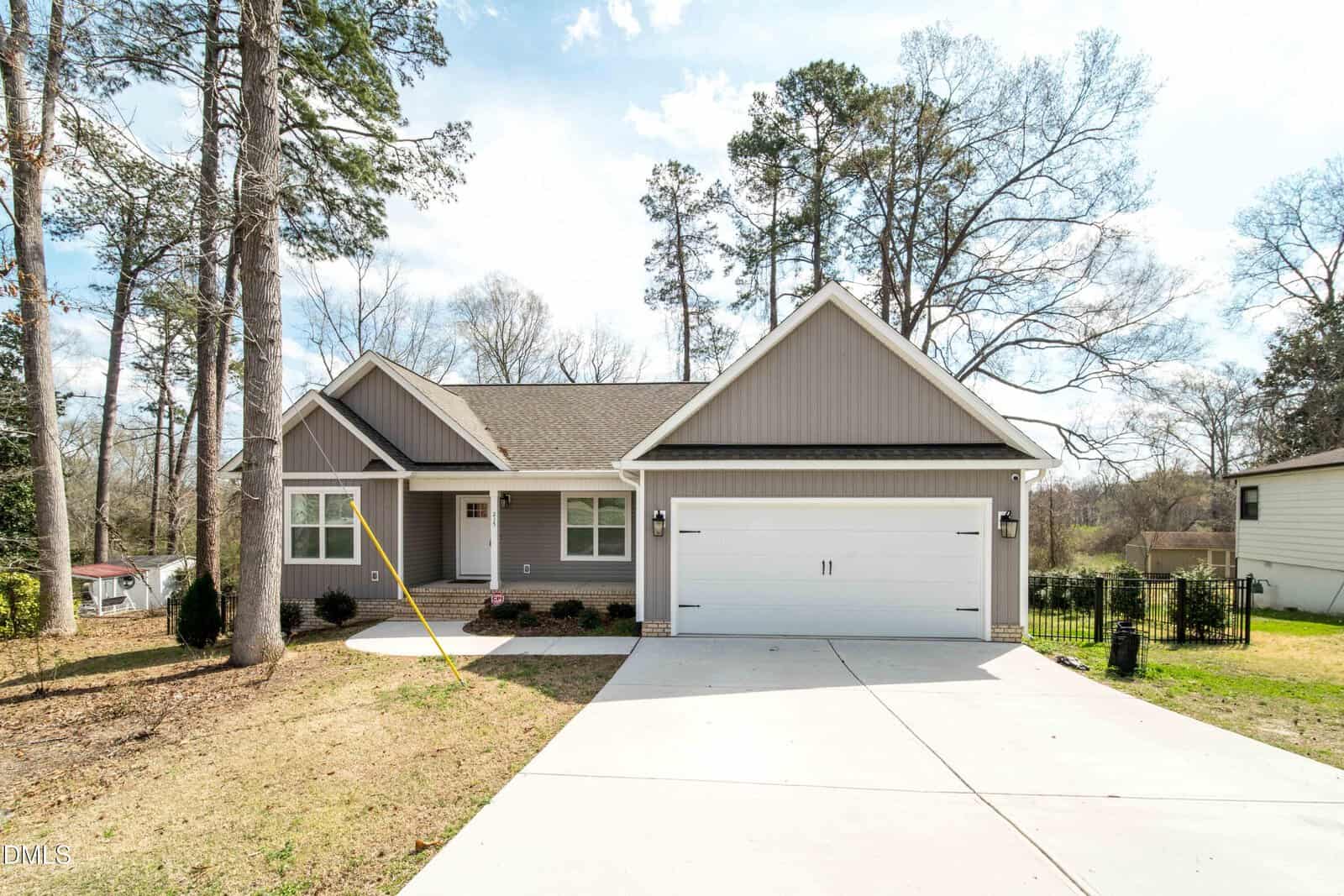 Single-story house with gray siding, white trim, and a two-car garage located at 215 Tanglewood Drive in Louisburg. The driveway is wide, and the yard is mostly dirt with sparse grass. Tall trees surround the area, and the sky is partly cloudy.