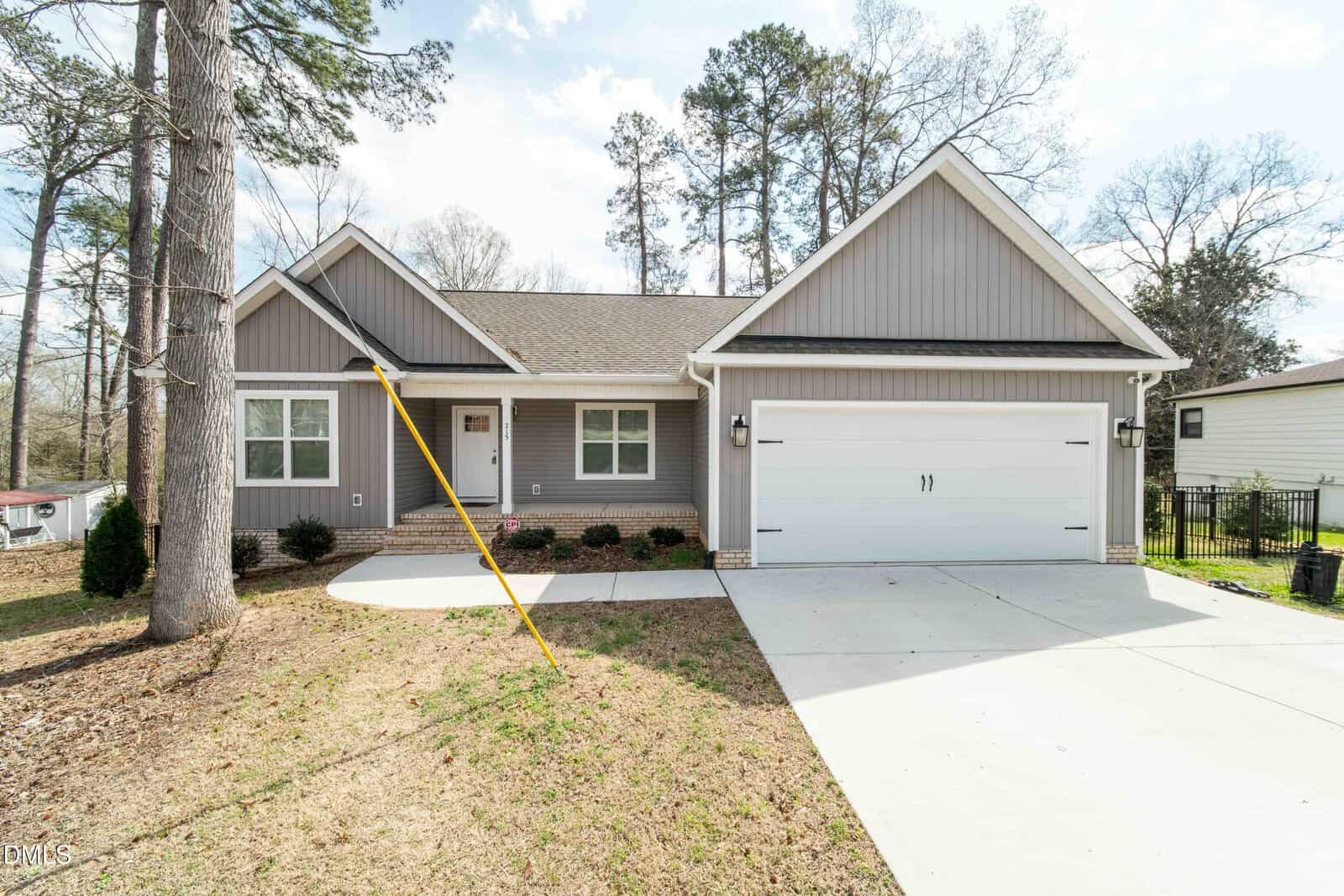 Single-story gray house with white trim and attached two-car garage, set on a sloped lot with a concrete driveway. Tall trees and a lawn surround the house. The front entrance has steps and small shrubs along the foundation.