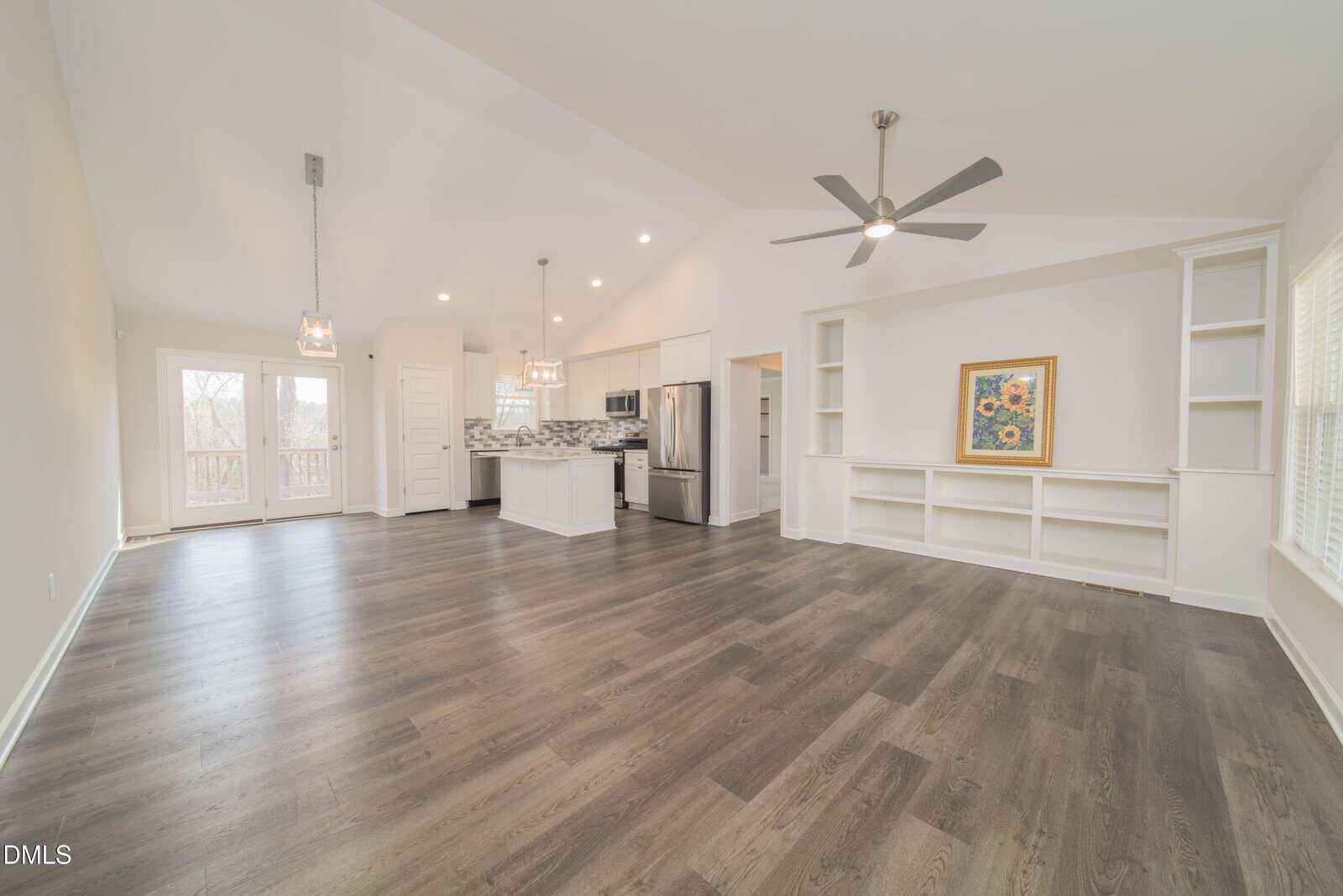 Bright open-concept living area with wood floors, ceiling fan, built-in shelves, and large windows. The modern kitchen features white cabinets, stainless steel appliances, and pendant lights over an island.