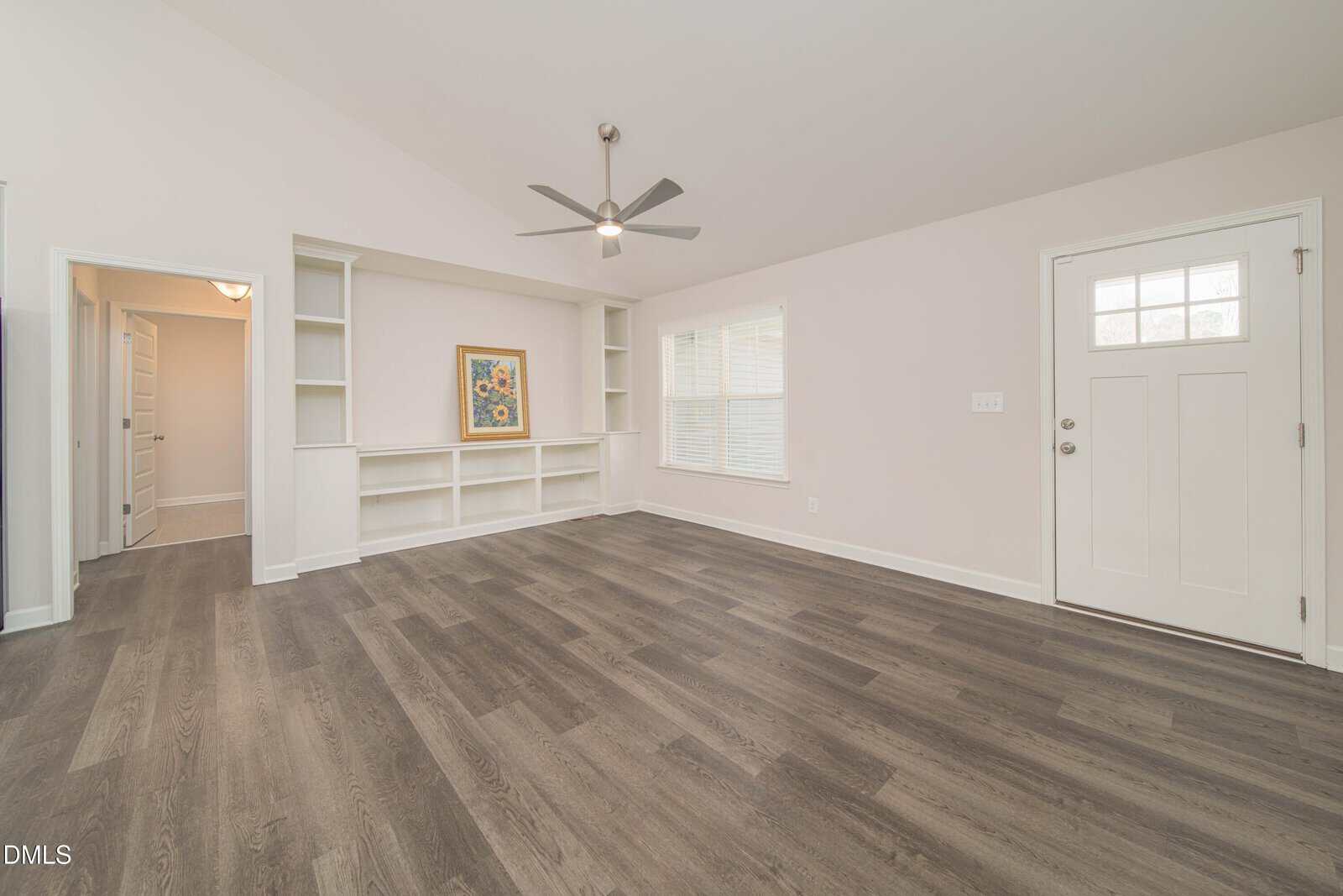 Bright, empty living room with light walls, dark wood flooring, ceiling fan, built-in shelves, a framed picture, two windows, and a white front door. An open doorway leads to another room.
