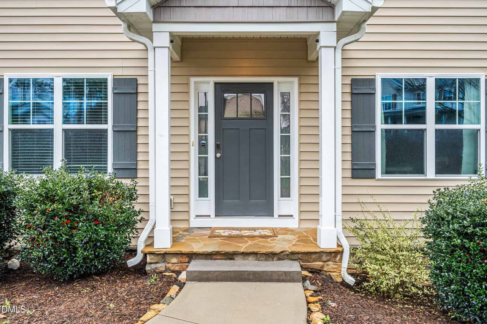 Front entrance of a beige house with gray front door, two side windows, a stone step, welcome mat, and shrubs lining the walkway leading to the porch.