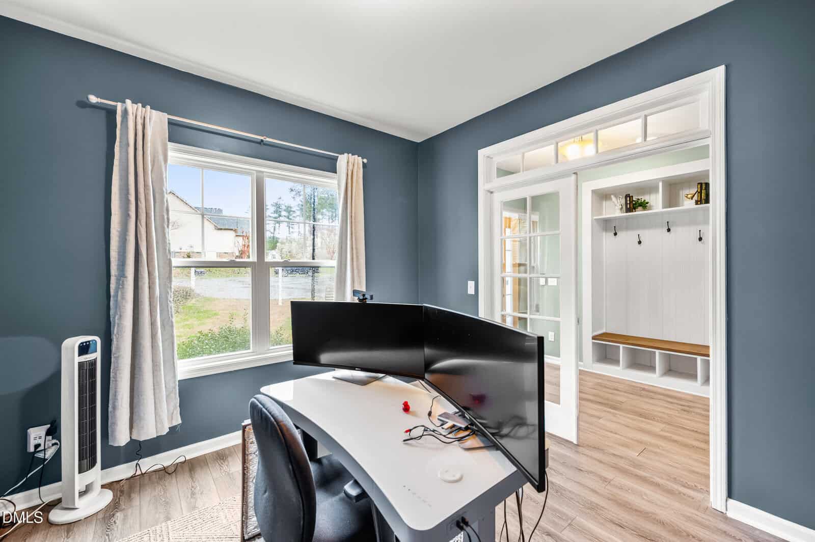 A home office with a curved monitor on a white desk, office chair, and fan by a window with beige curtains. Through double glass doors, a mudroom with hooks and a wooden bench is visible. Walls are painted dark blue.