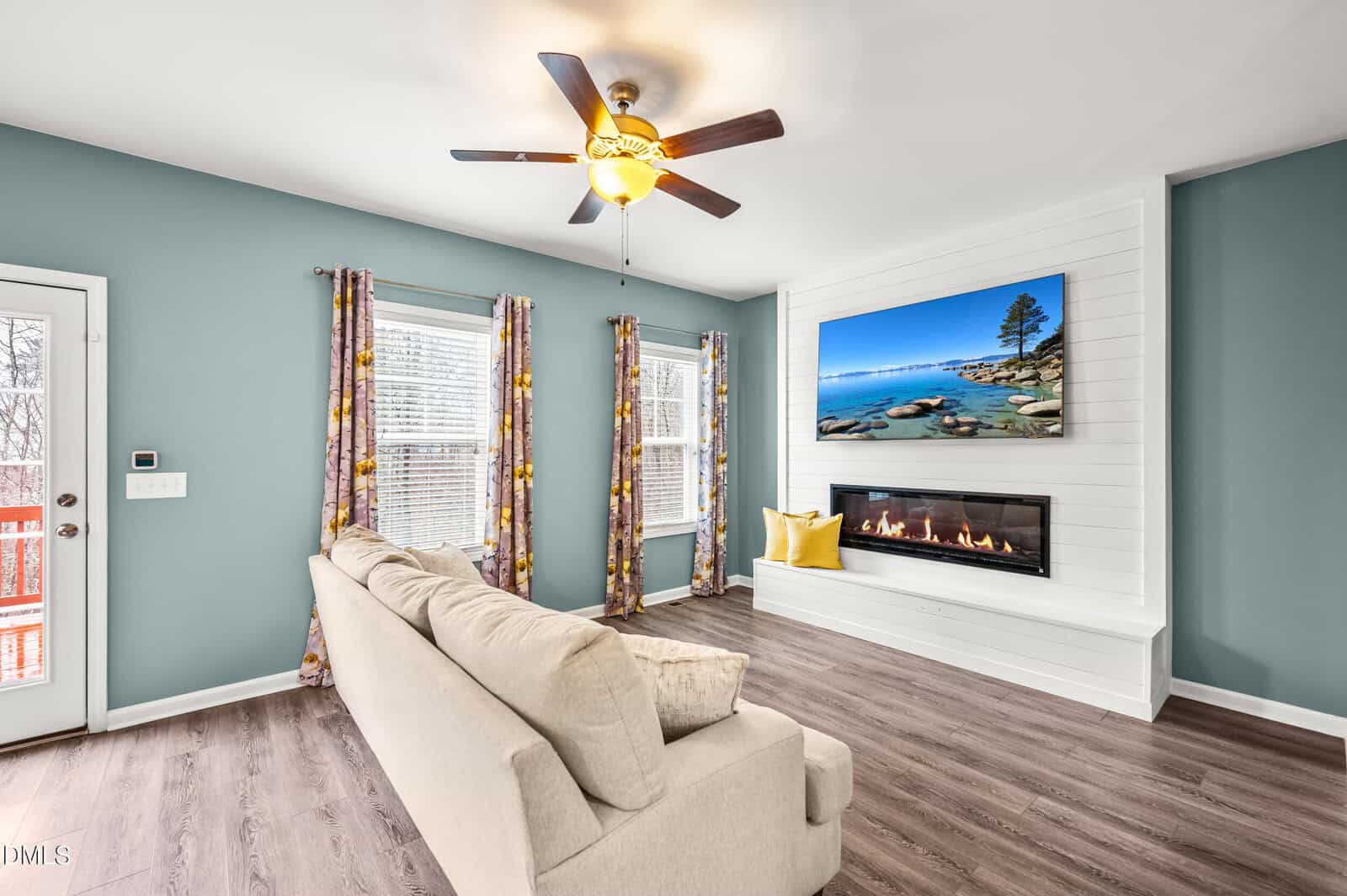 A living room with light blue walls, a beige sofa, floral curtains, wood flooring, a ceiling fan with a light, and a built-in fireplace beneath a wall-mounted TV displaying a nature scene.