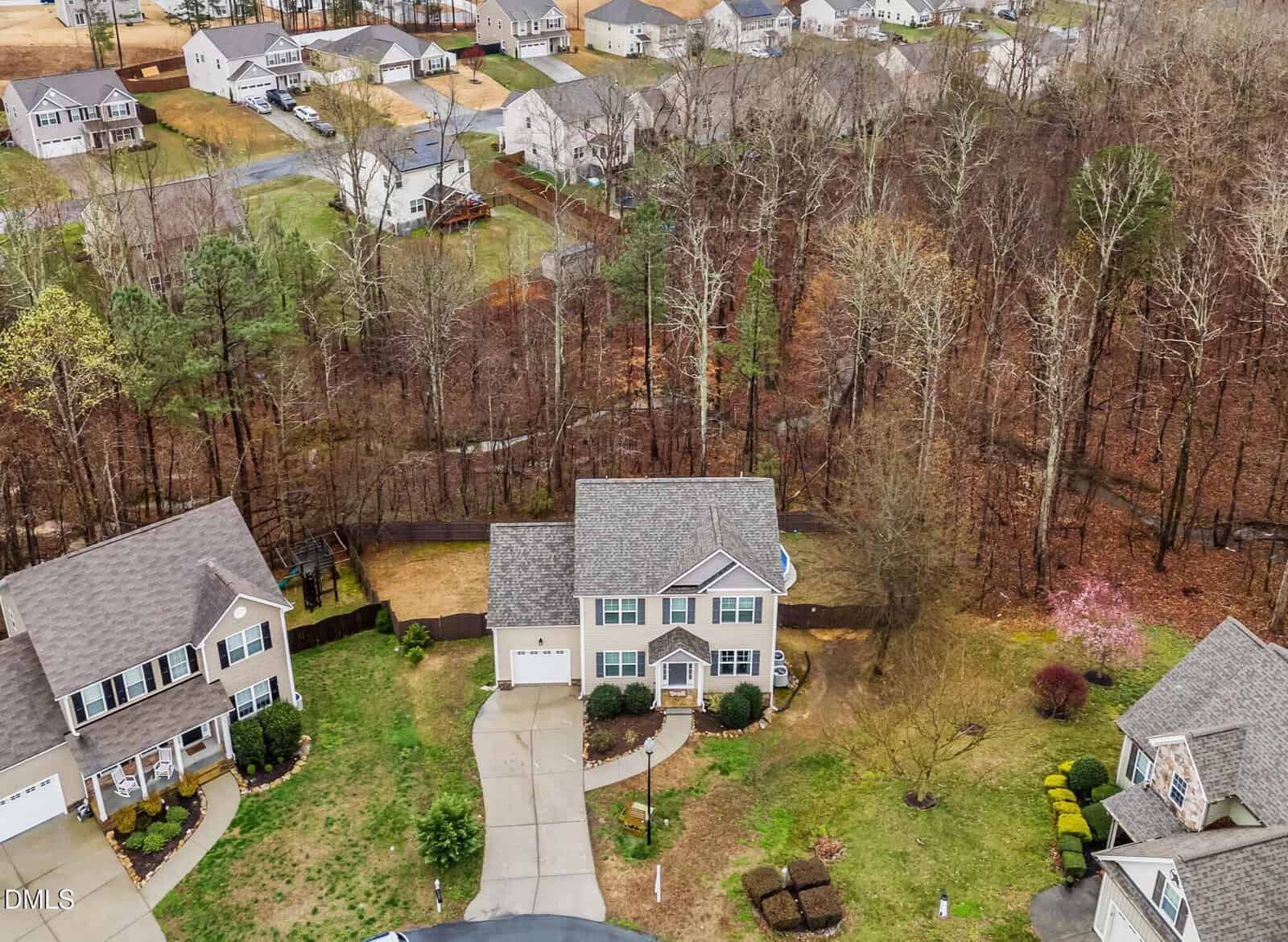 Aerial view of a suburban neighborhood with several houses, driveways, and yards adjacent to a wooded area with leafless trees. The central house has a large backyard and is bordered by a curved driveway.