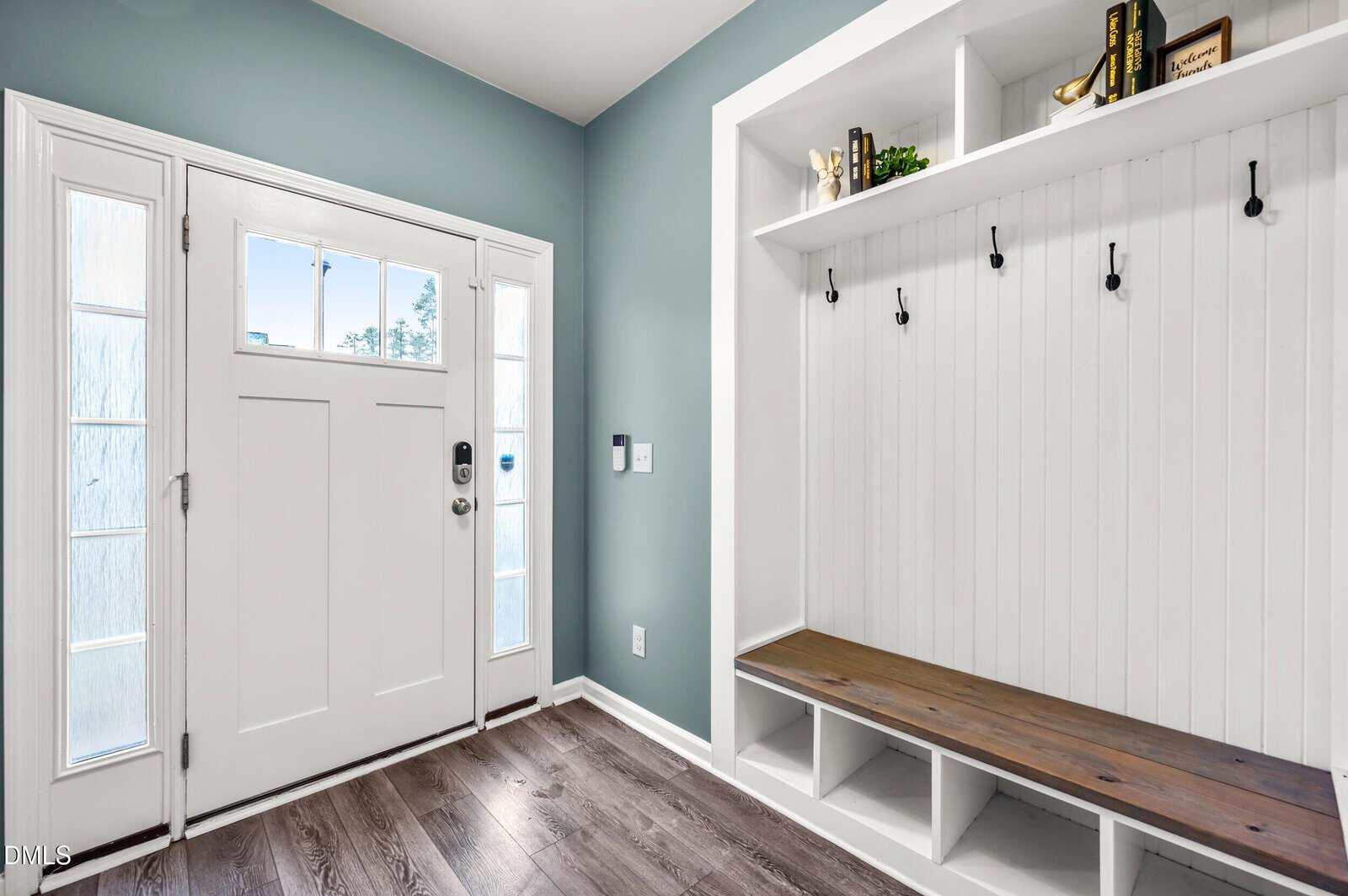 Bright entryway with a white front door, side windows, blue-green walls, and a built-in white bench featuring hooks and cubbies for storage. Decor and books are placed on the top shelf above the hooks.