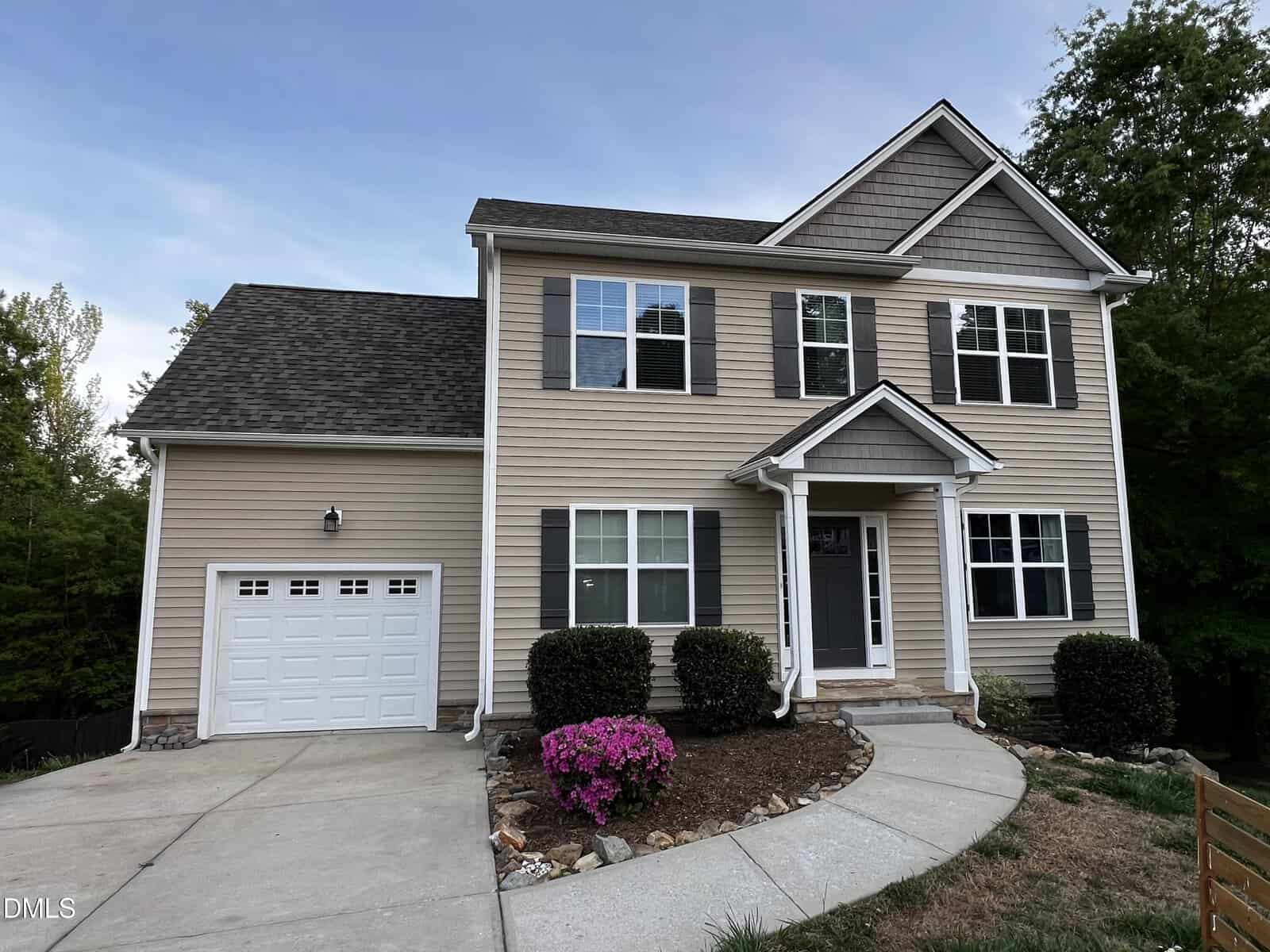 Charming two-story beige house at 102 Griffis Drive, Stem, with white trim, gray shutters, and a one-car garage. Enjoy the cozy covered porch, landscaped yard with bushes and blooming pink flower, plus a curved driveway leading to the front door.