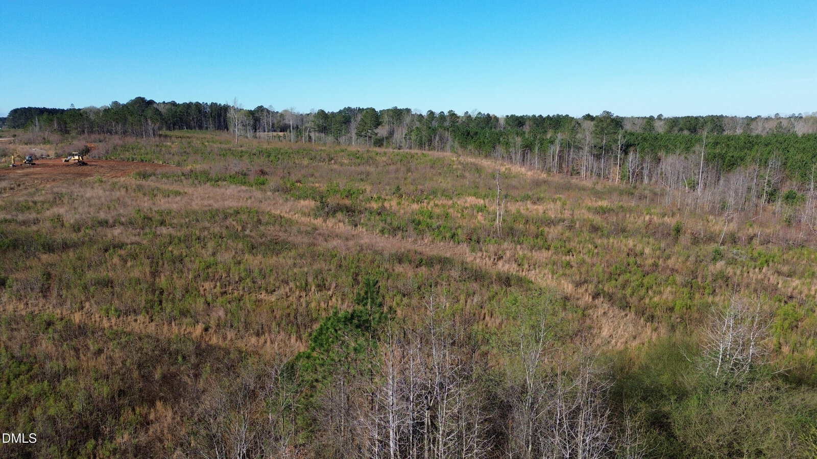 A large, open field with patches of grass and small shrubs, bordered by a line of trees under a clear blue sky. Construction equipment is visible in the distance on the left side.